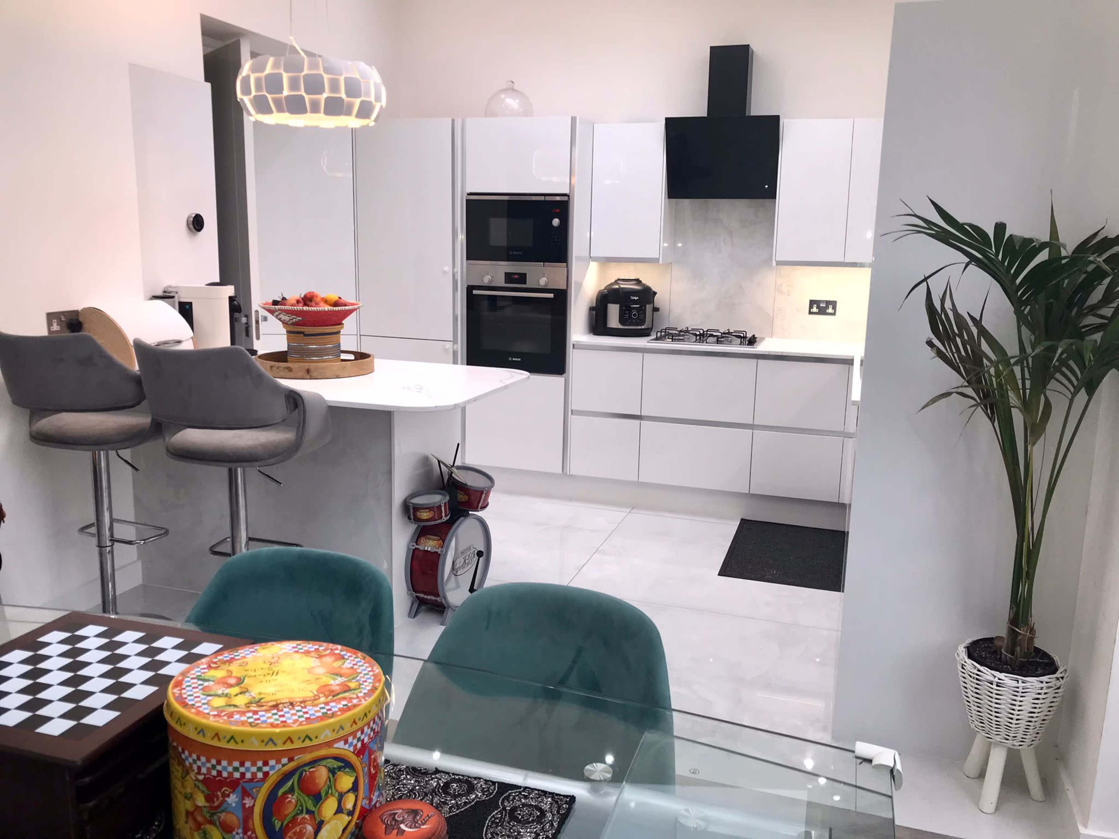 A modern kitchen with white cabinetry, sleek appliances, a dining area featuring a glass table, and decorative items like a checkered board and colorful containers.