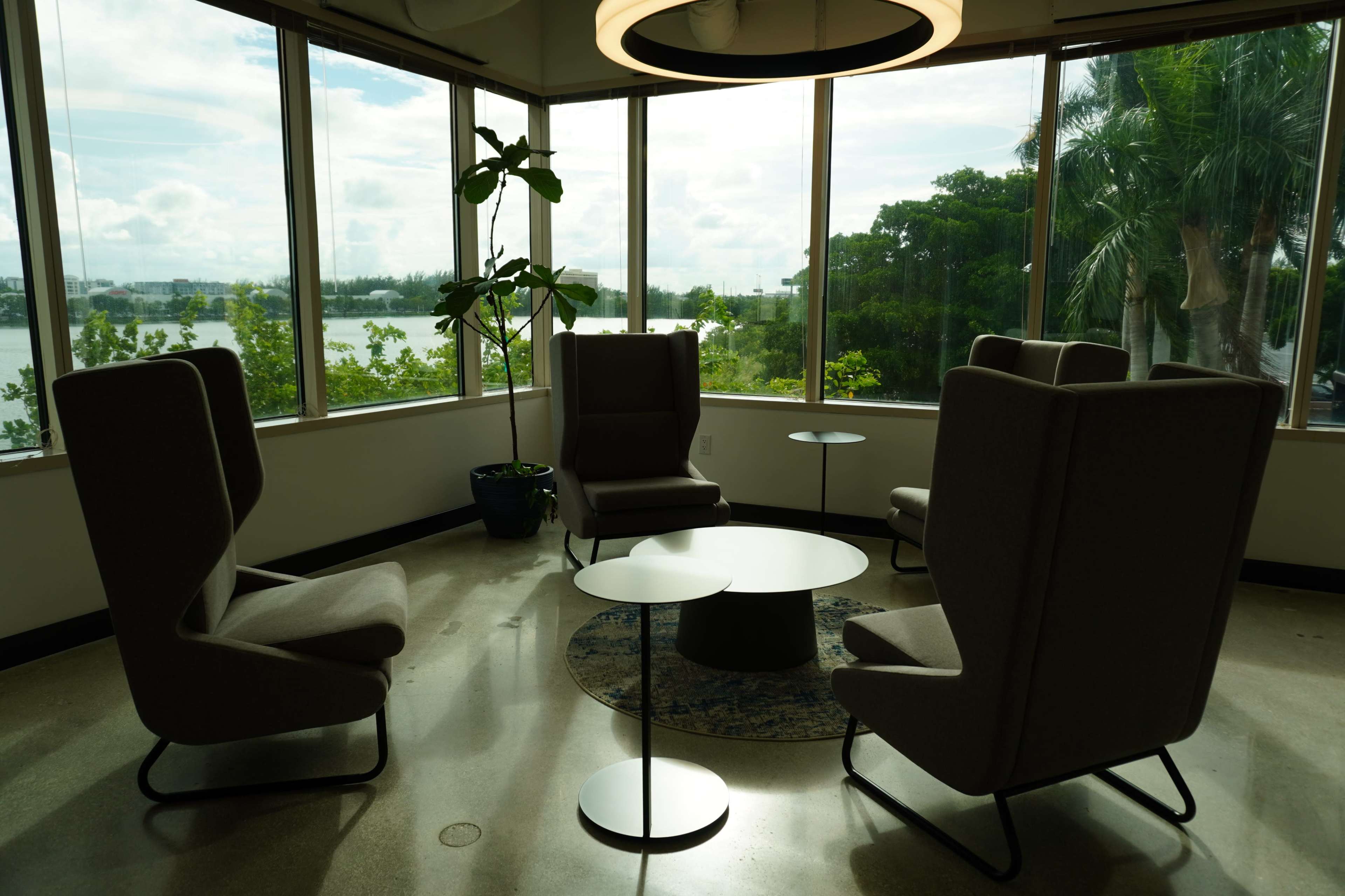 The image shows a modern seating area with four high-backed chairs and a low coffee table, surrounded by large windows that provide a view of greenery and water outside.