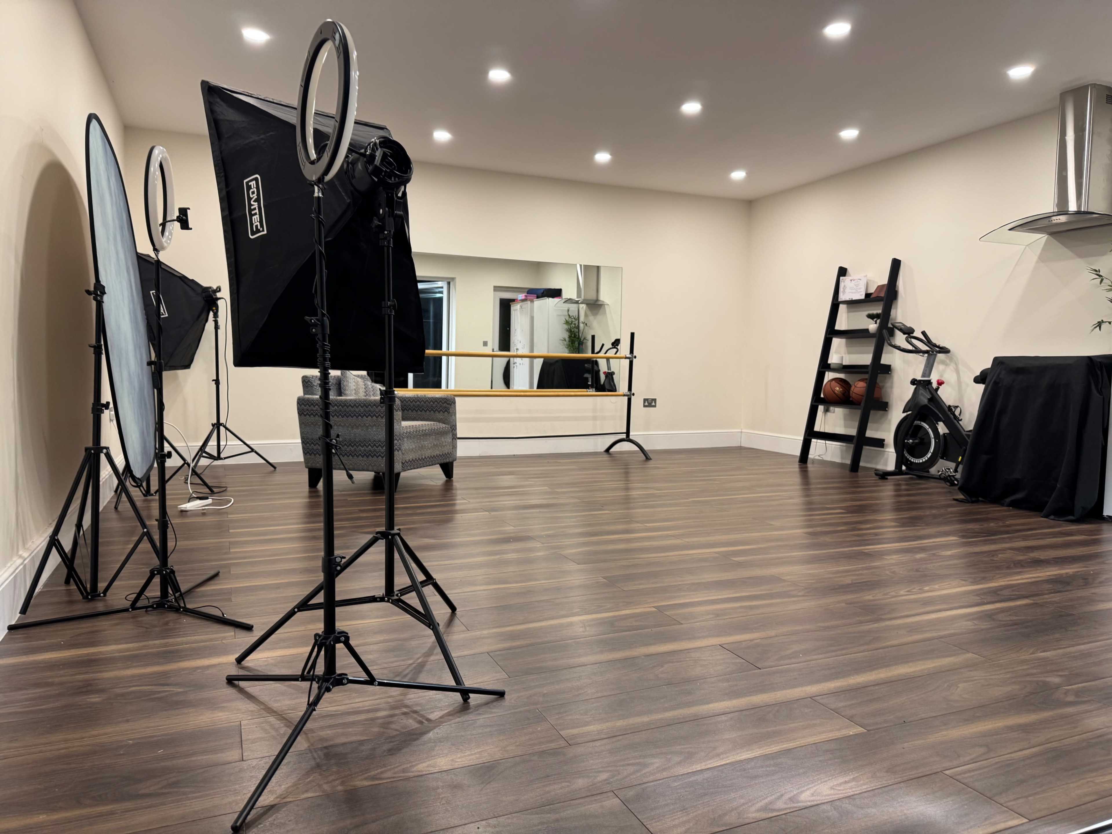 A spacious studio setting features professional lighting equipment, a mirror, a ballet bar, and exercise equipment in a well-lit room with wooden flooring.