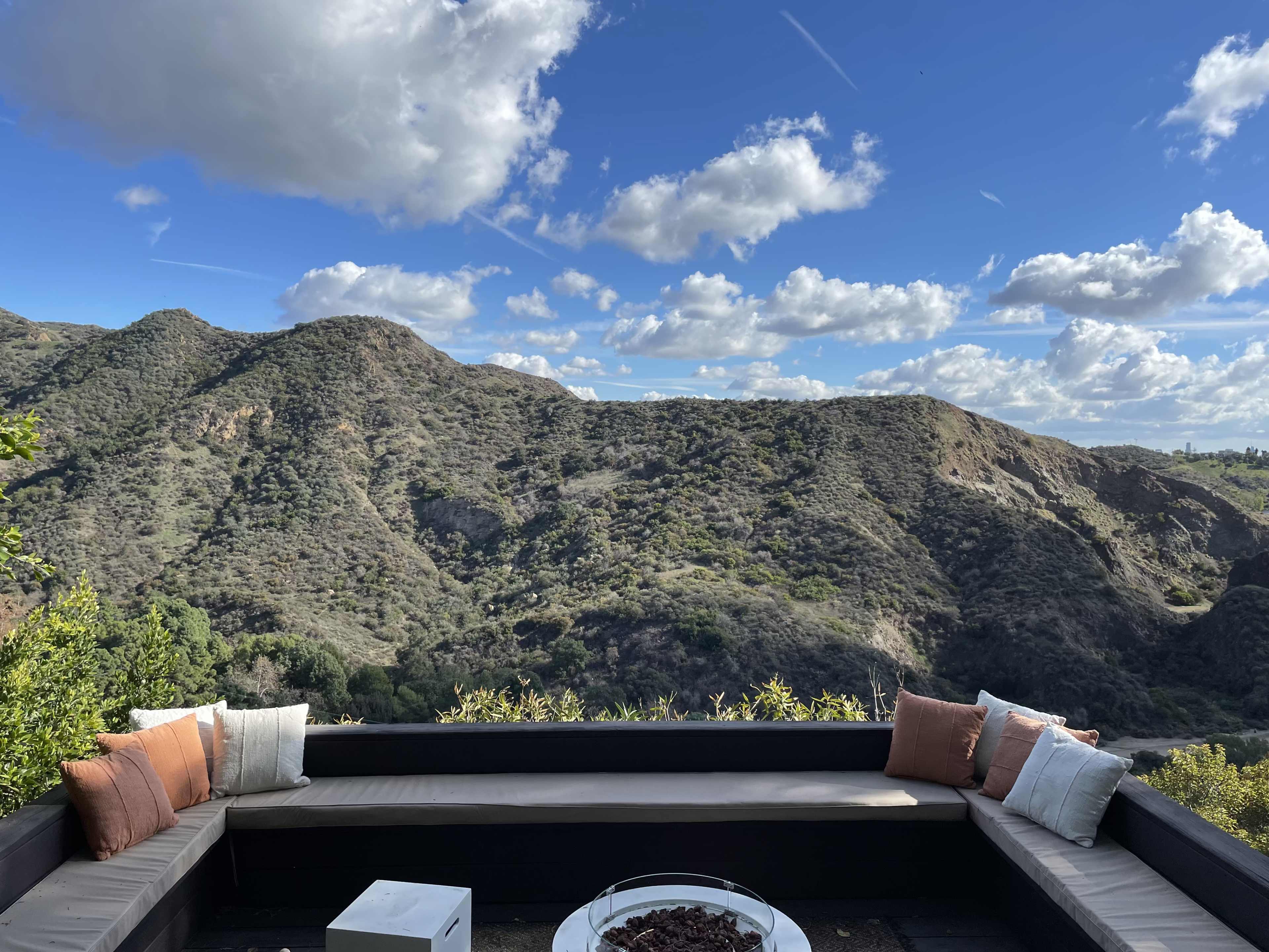 A seating area with cushions overlooking a hilly landscape under a partly cloudy sky.