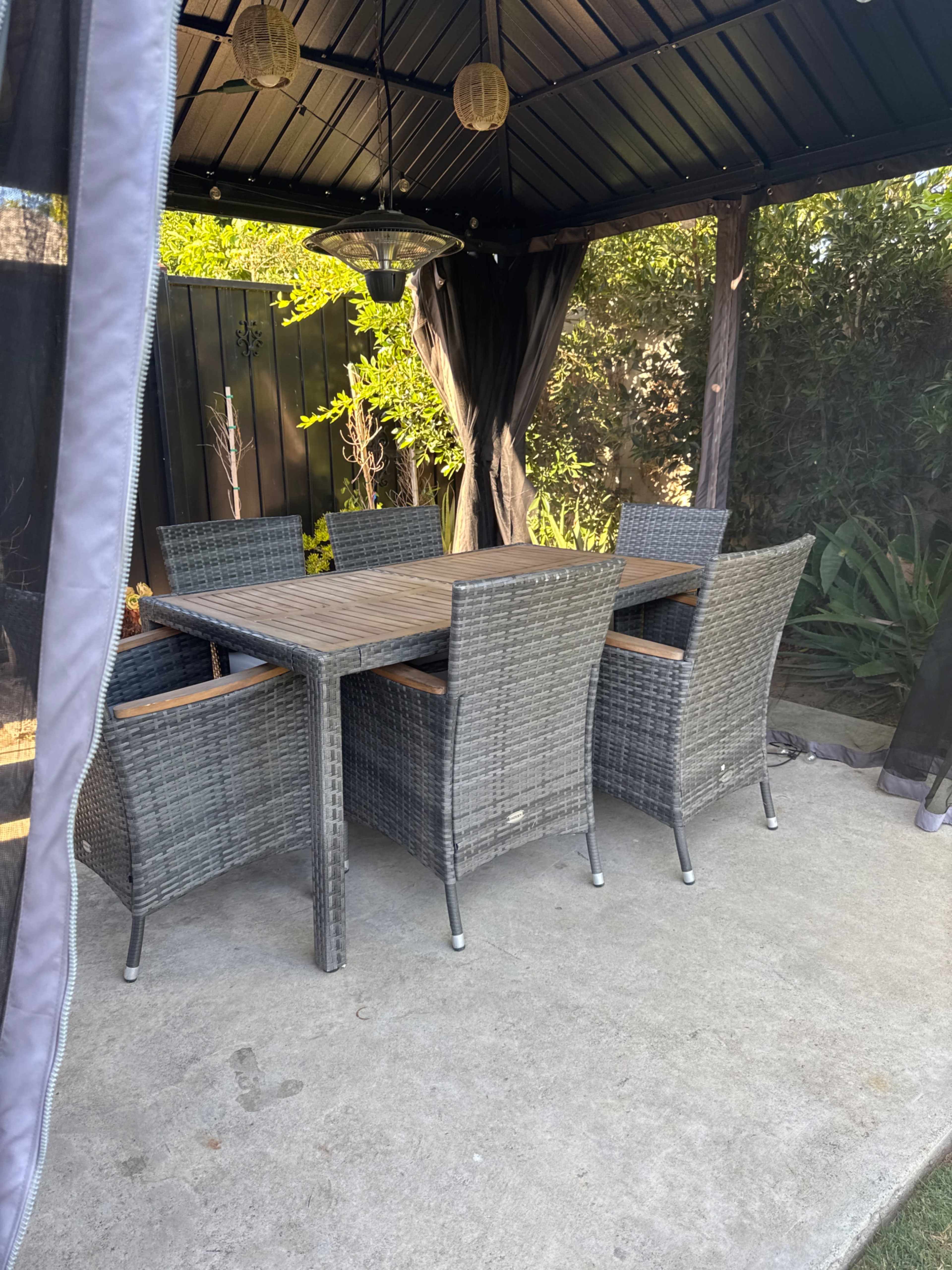 A set of six wicker dining chairs surrounds a rectangular table under a gazebo with greenery in the background.