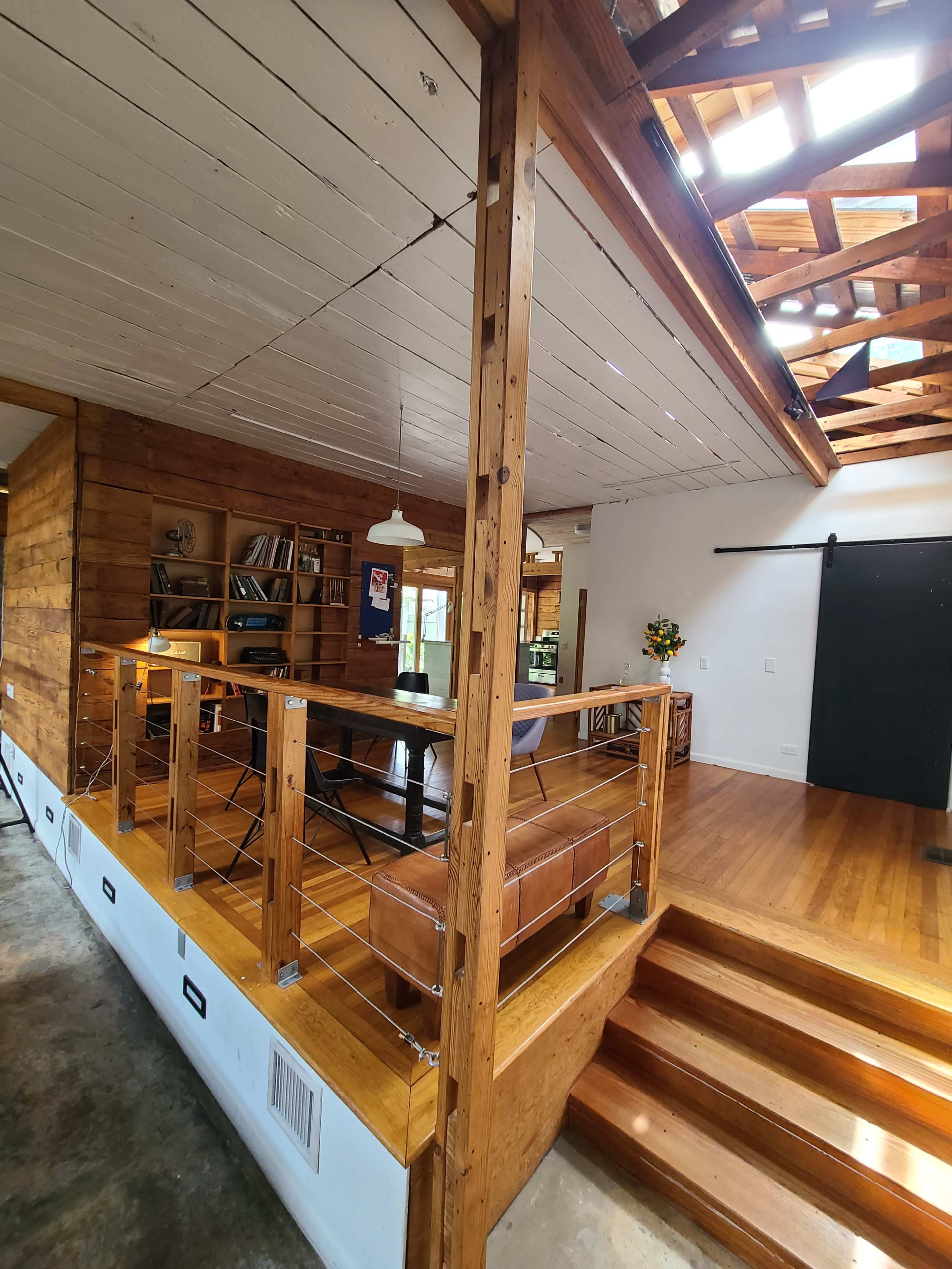 The interior of a wooden house features an open layout with a raised area that includes a lounge and shelves, illuminated by natural light from the ceiling.