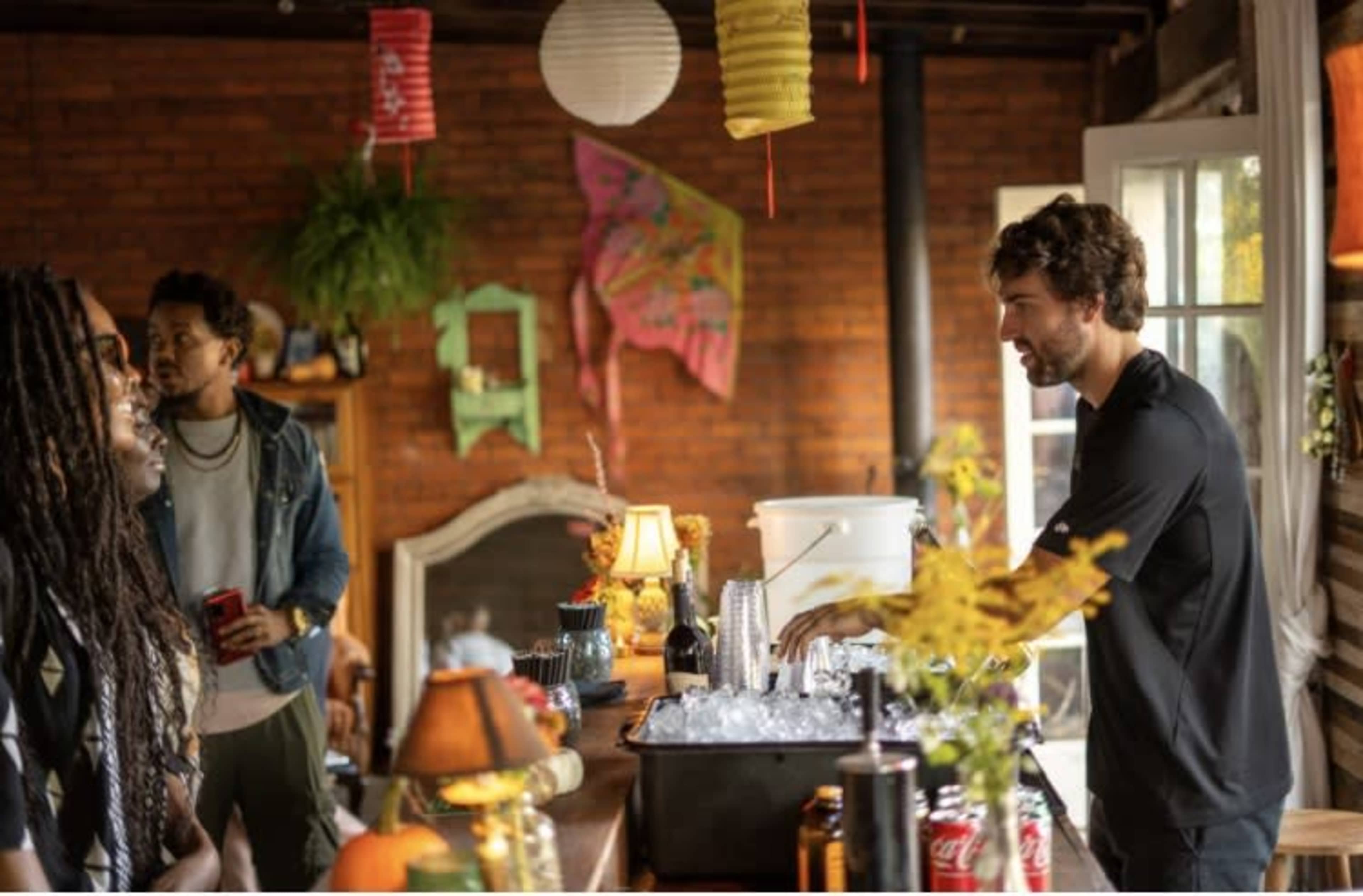 A group of three people conversations in a rustic room with a bar setup featuring drinks and decorative lanterns.