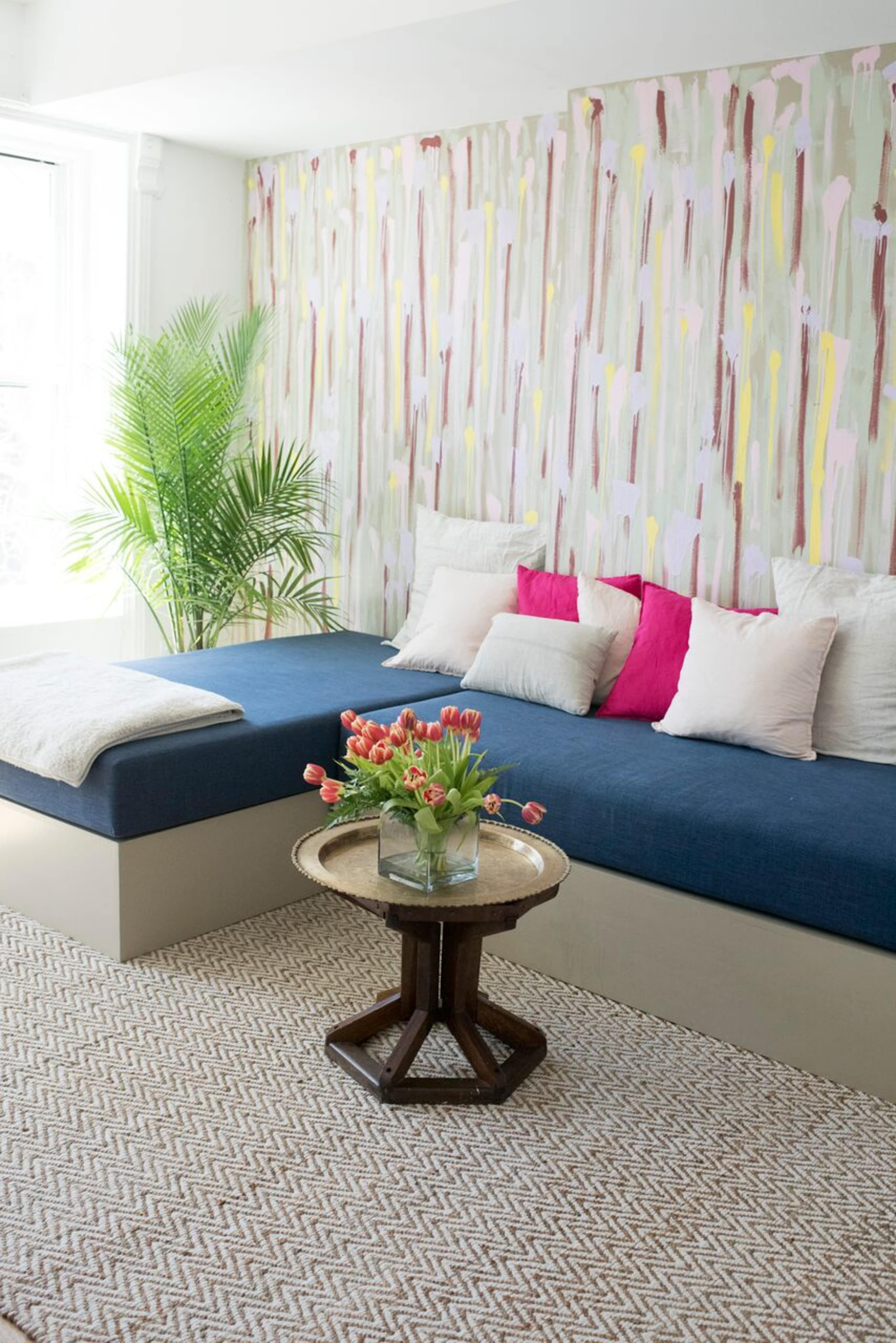 A modern living space with a blue sofa adorned with cushions, a round wooden coffee table topped with a vase of tulips, and a decorative wall with a colorful, abstract design.