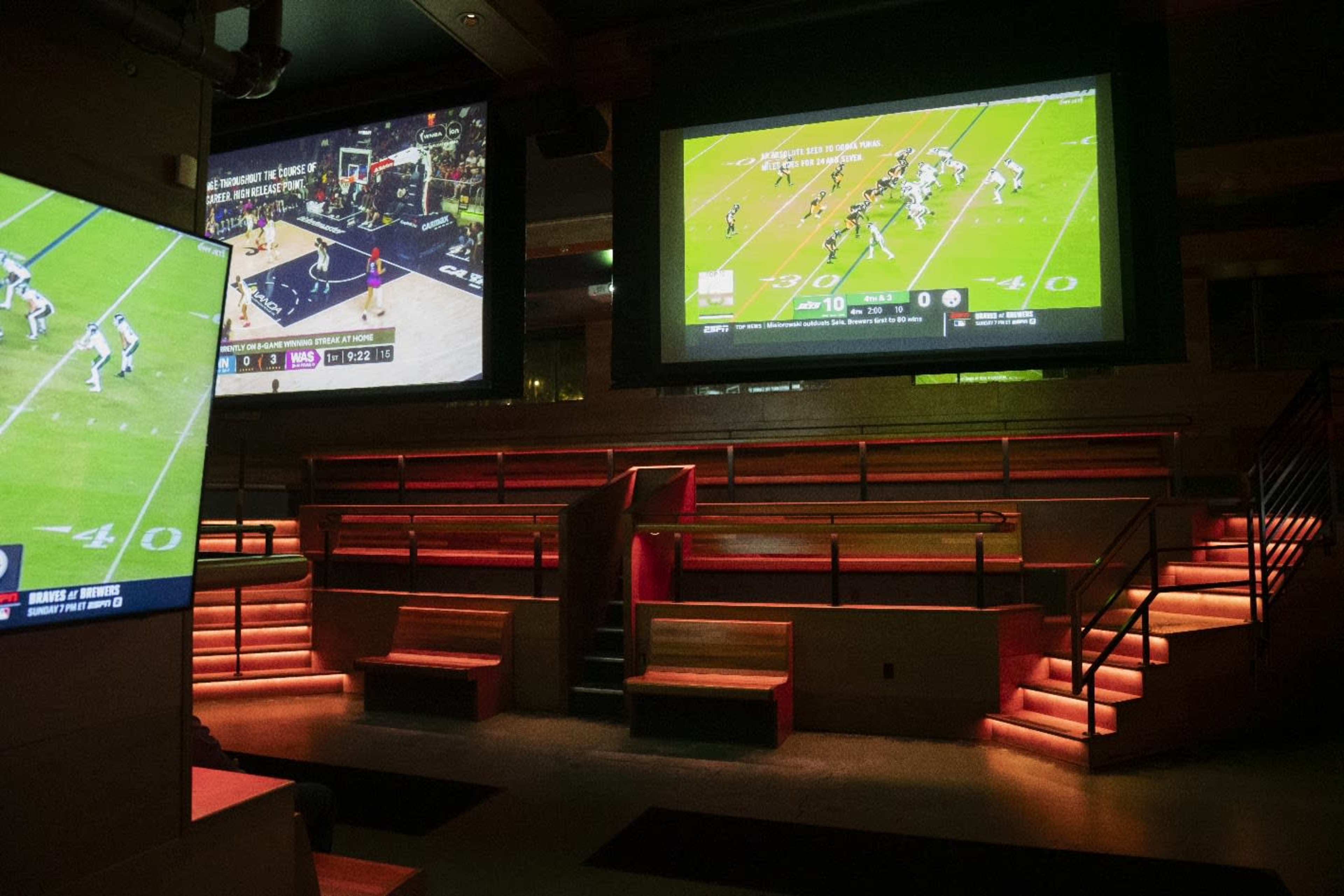 The image shows a modern sports bar with multiple large screens displaying live sports games and seating arranged in a tiered layout.
