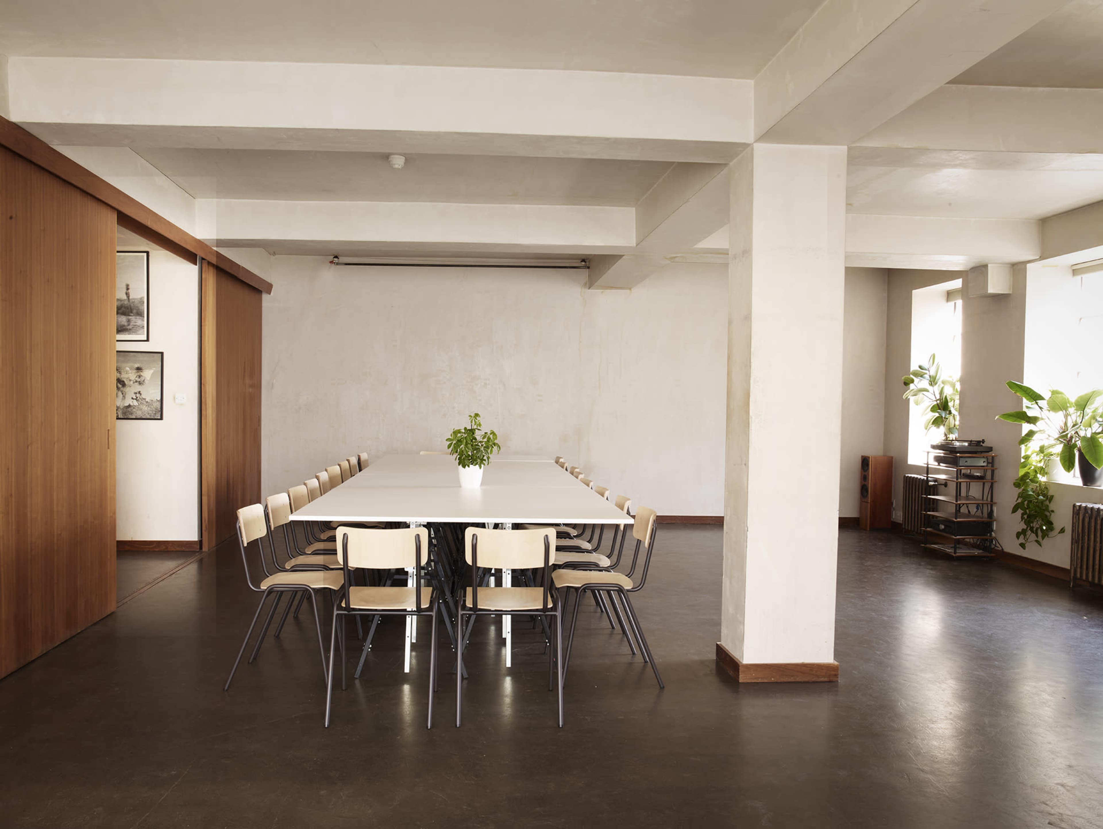 A large, empty conference table surrounded by wooden chairs is set in a spacious, minimalist room with plants and wooden accents.