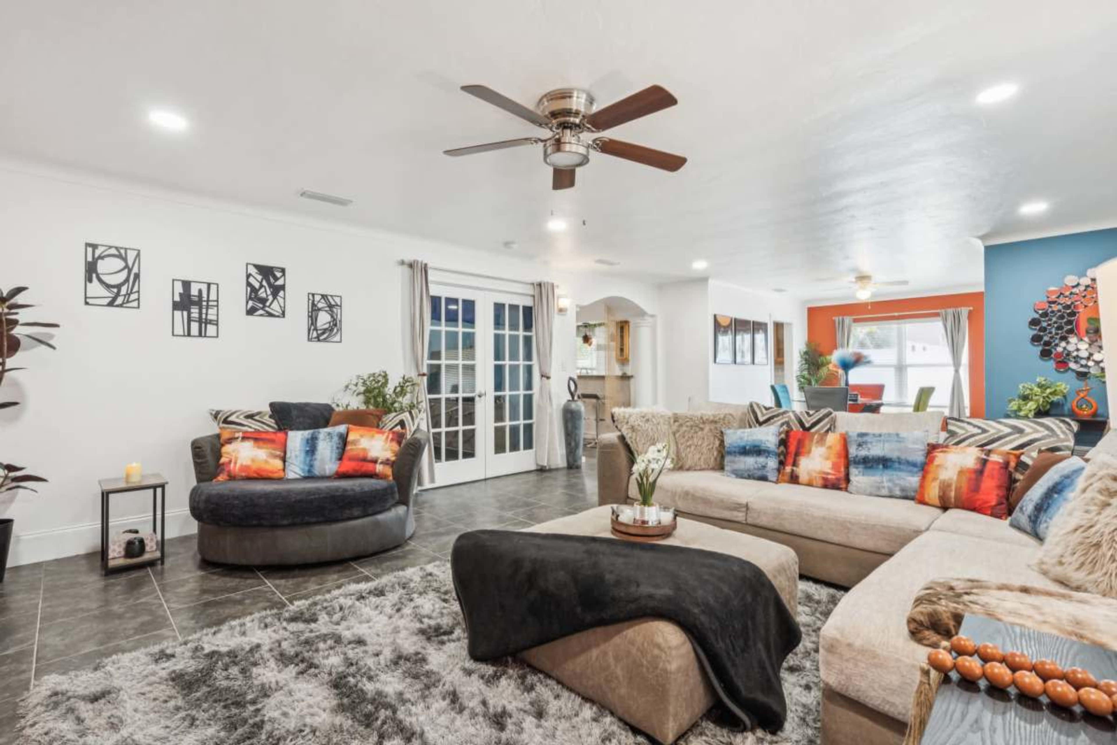 The image shows a spacious living room featuring a sectional sofa, a circular chair, a large rug, and colorful decorative cushions on a tiled floor.