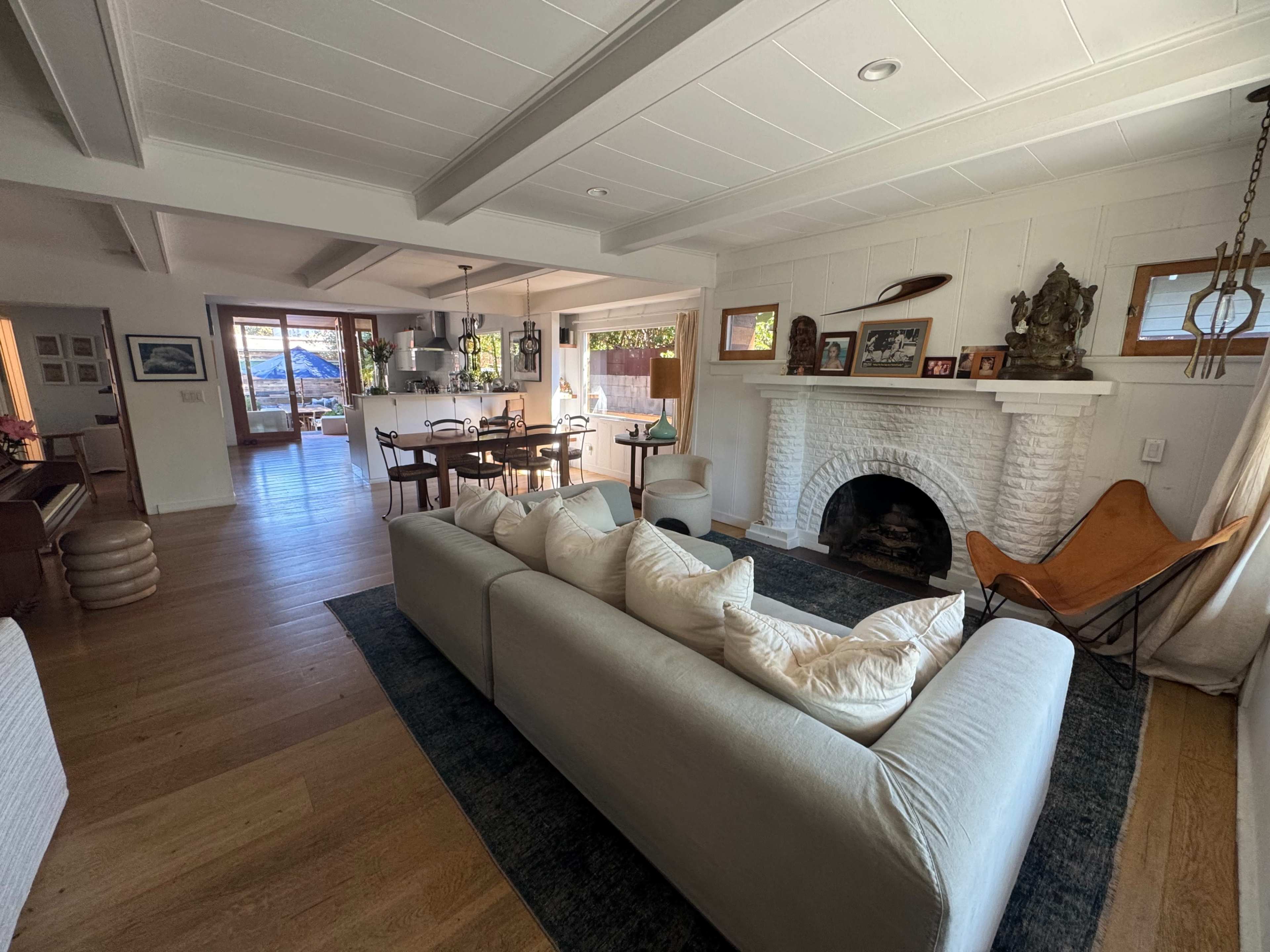 The image displays a spacious living room with a white brick fireplace, adjacent to a dining area and a kitchen, featuring wooden floors and large windows.
