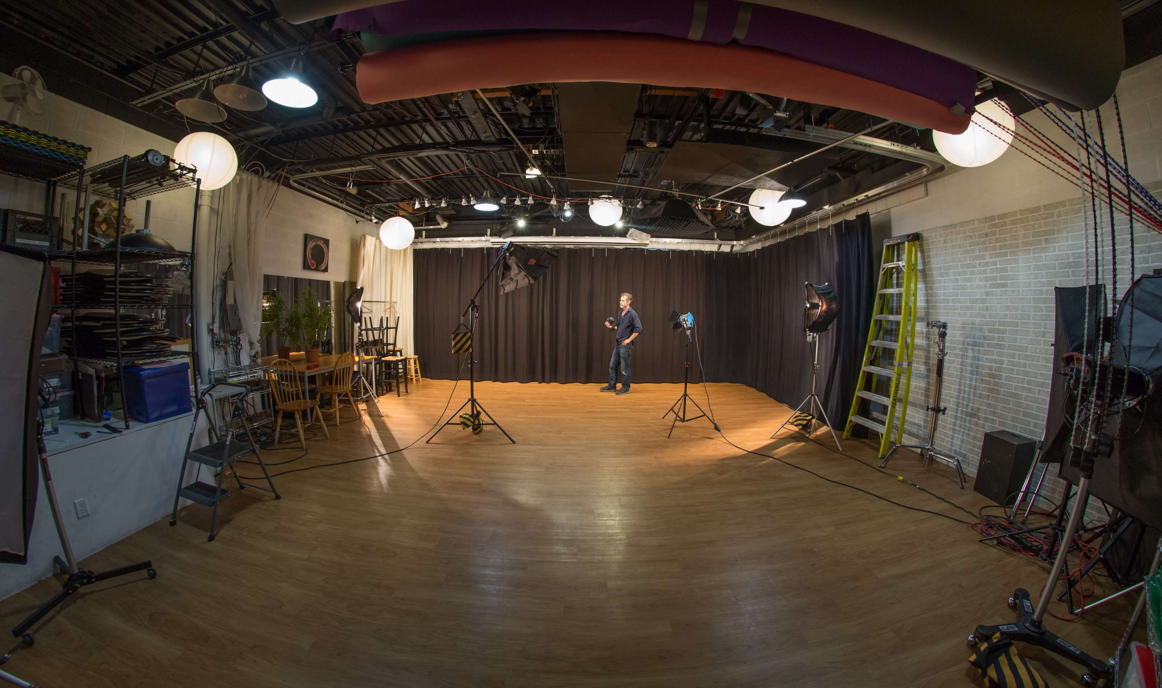 A photography studio with a wooden floor, black curtains, and various lighting equipment set up around a central area.