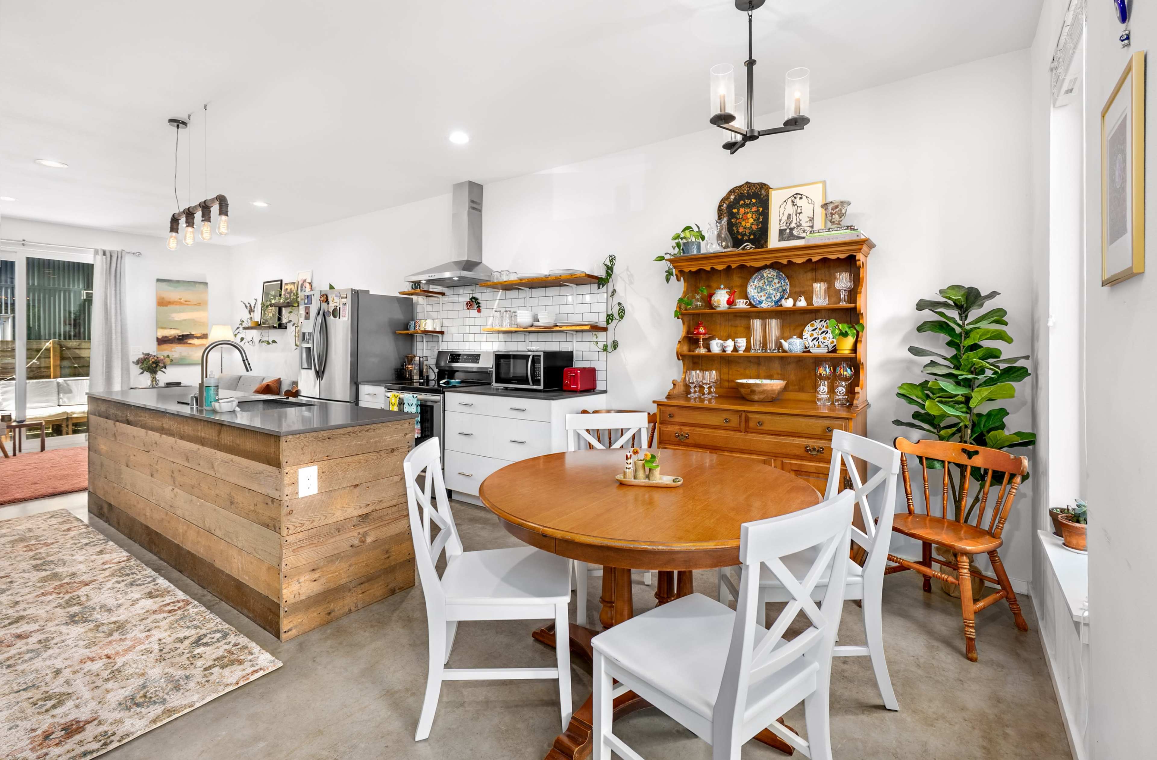 The image shows a modern kitchen with a wooden dining table surrounded by white chairs, a rustic island, and various kitchen appliances.