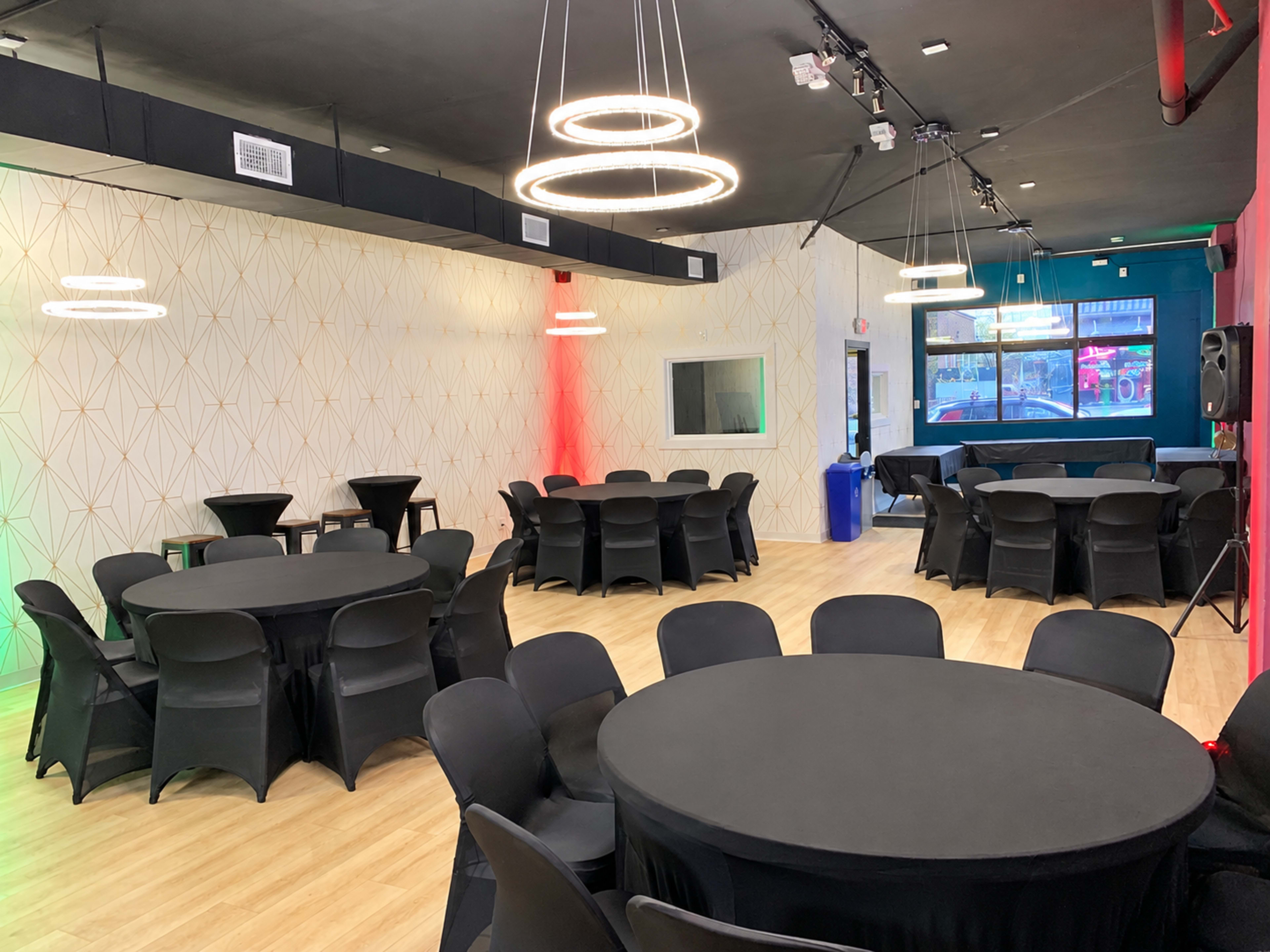 The image shows a spacious event room filled with round tables covered in black tablecloths, illuminated by modern pendant lights on the ceiling.