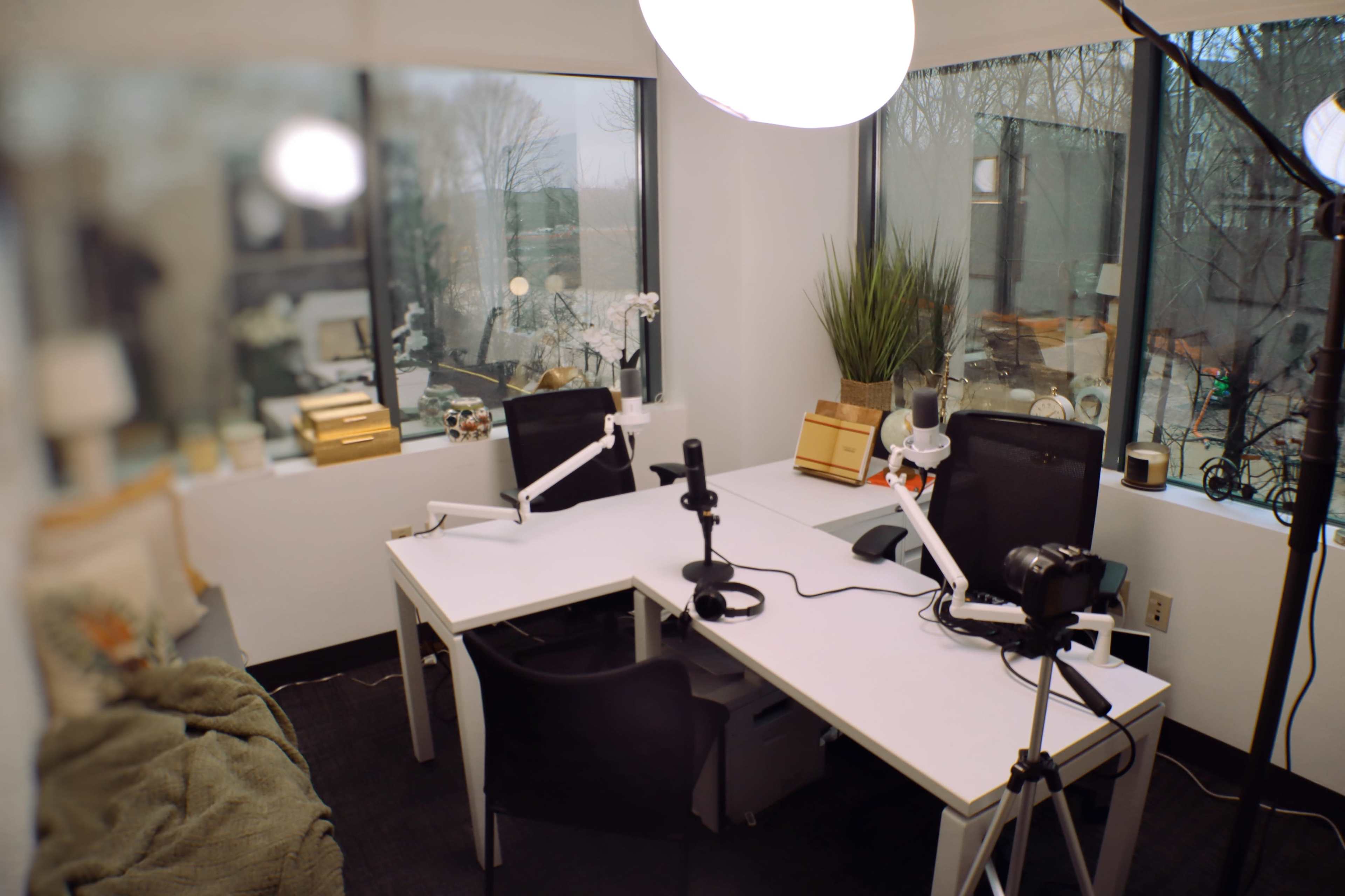 A modern office workspace features two desks with microphones and cameras set up, surrounded by large windows overlooking a natural landscape.
