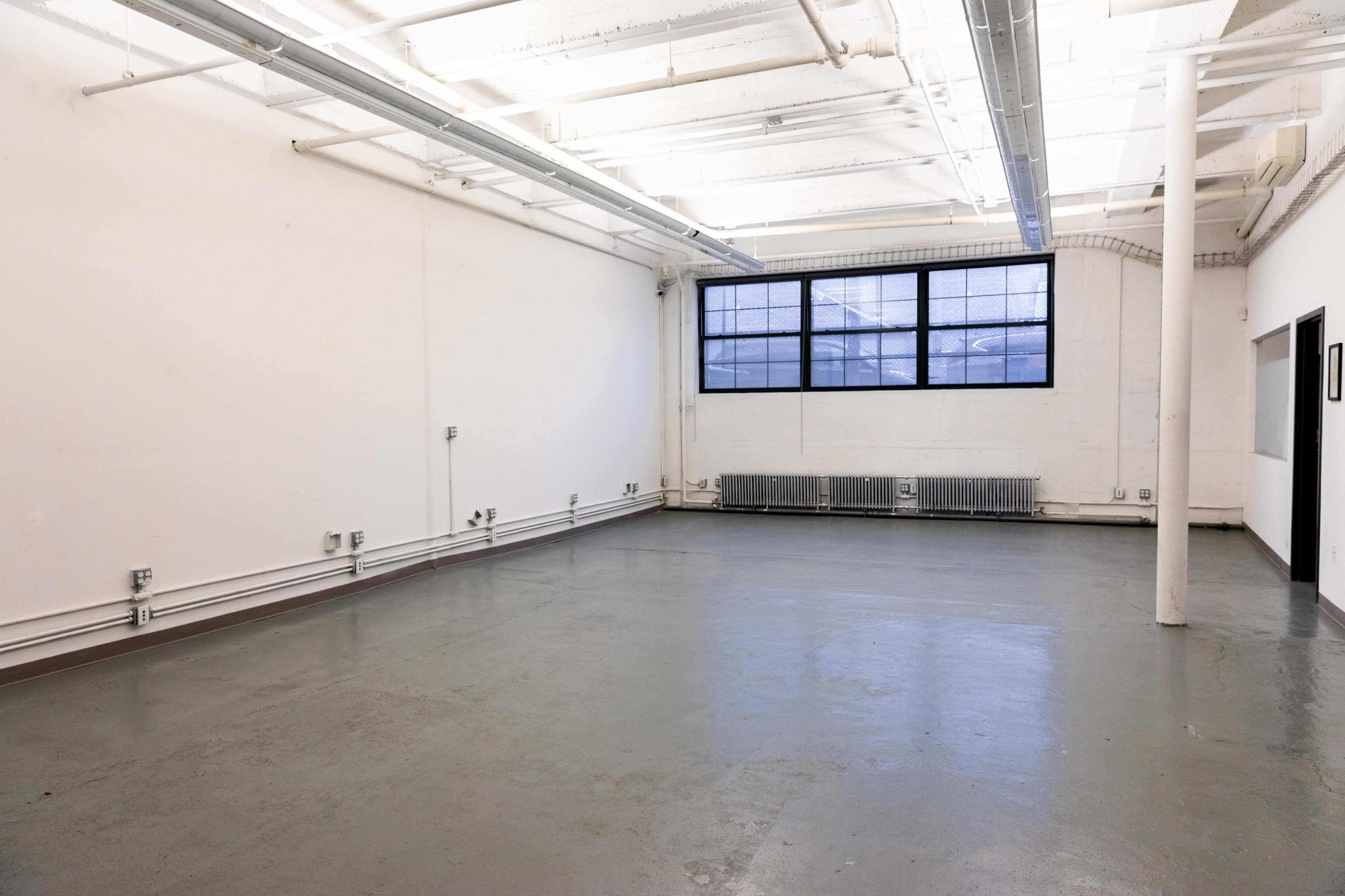 The image shows an empty industrial-style room with a concrete floor, exposed ceiling beams, and a large window on one wall.
