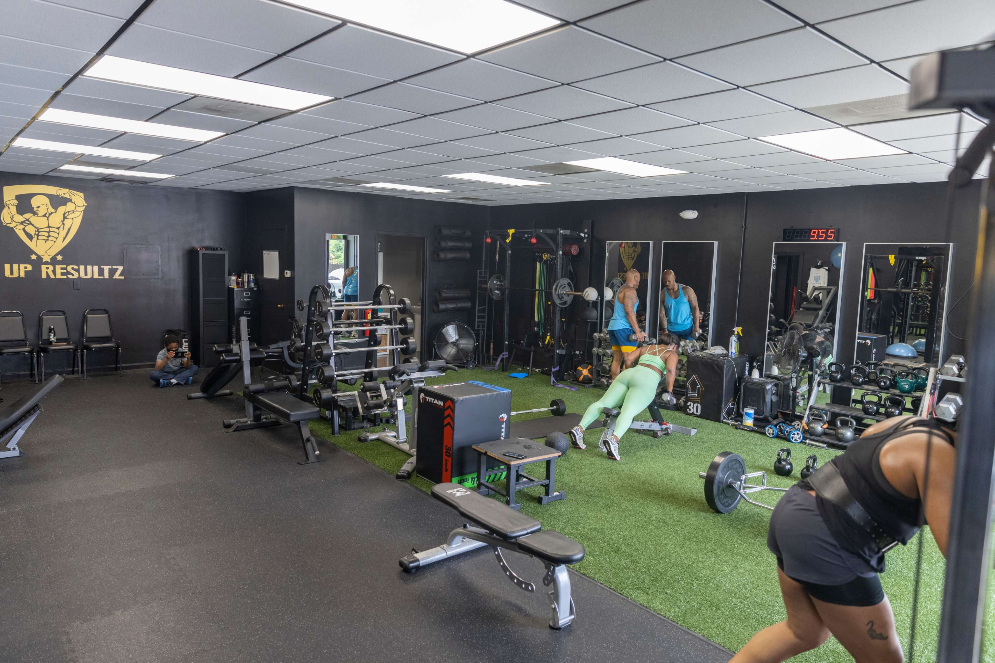 A gym interior with various exercise equipment, including weights and benches, and two individuals engaged in workouts on a green turf area.