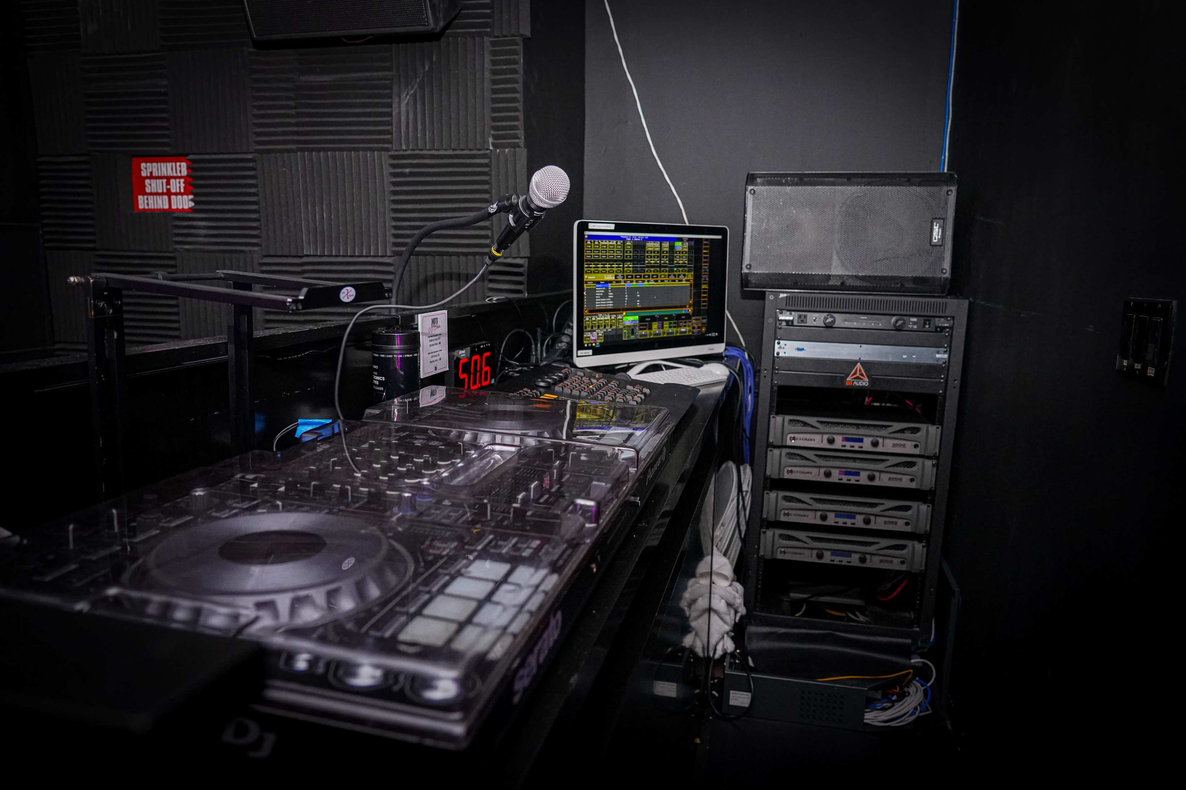 A DJ setup is shown, featuring a mixer, microphone, computer, and sound equipment in a darkened room.