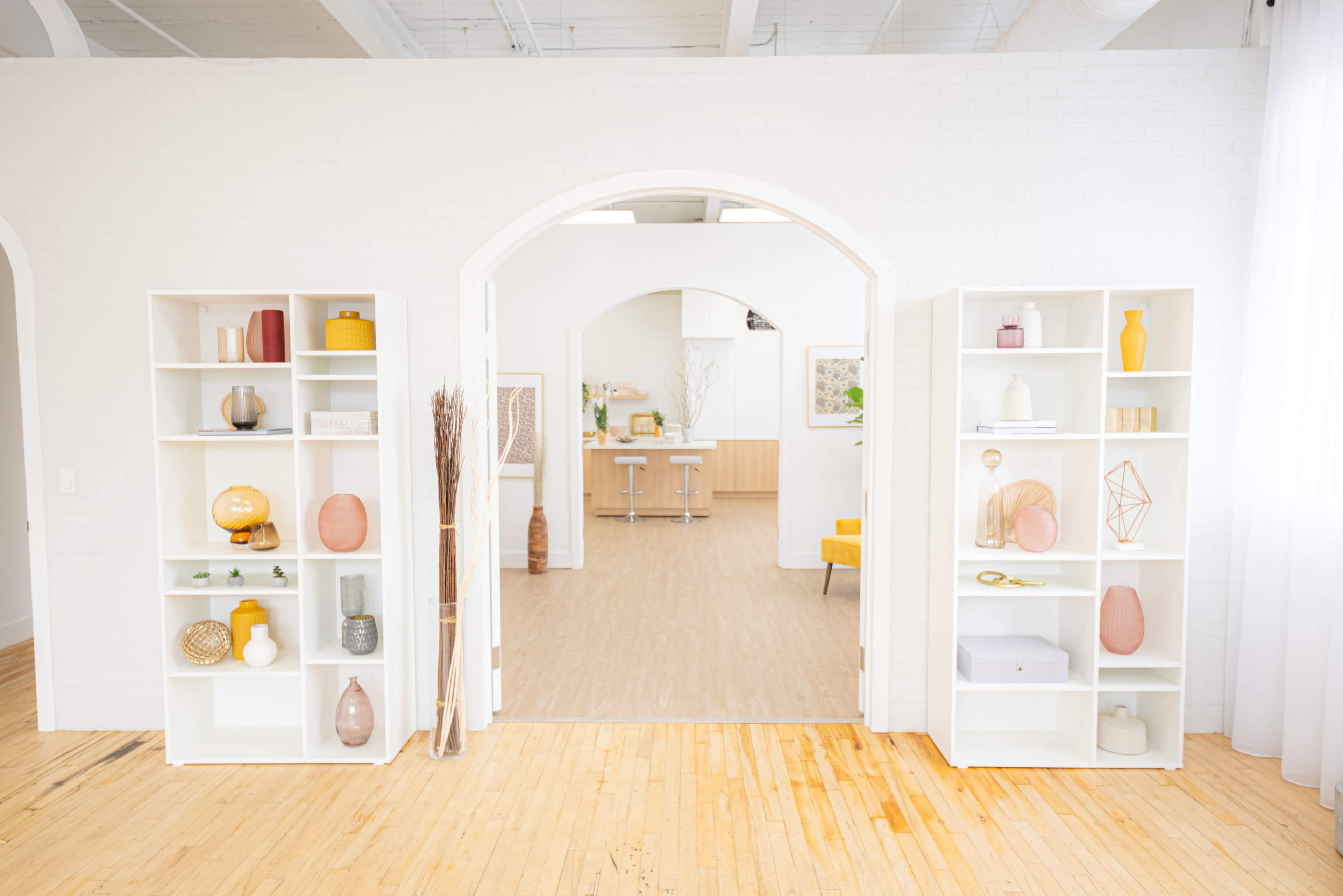 A bright room with two white bookshelves on either side of an open archway, leading to a light-furnished area with wooden flooring and minimalistic decor.