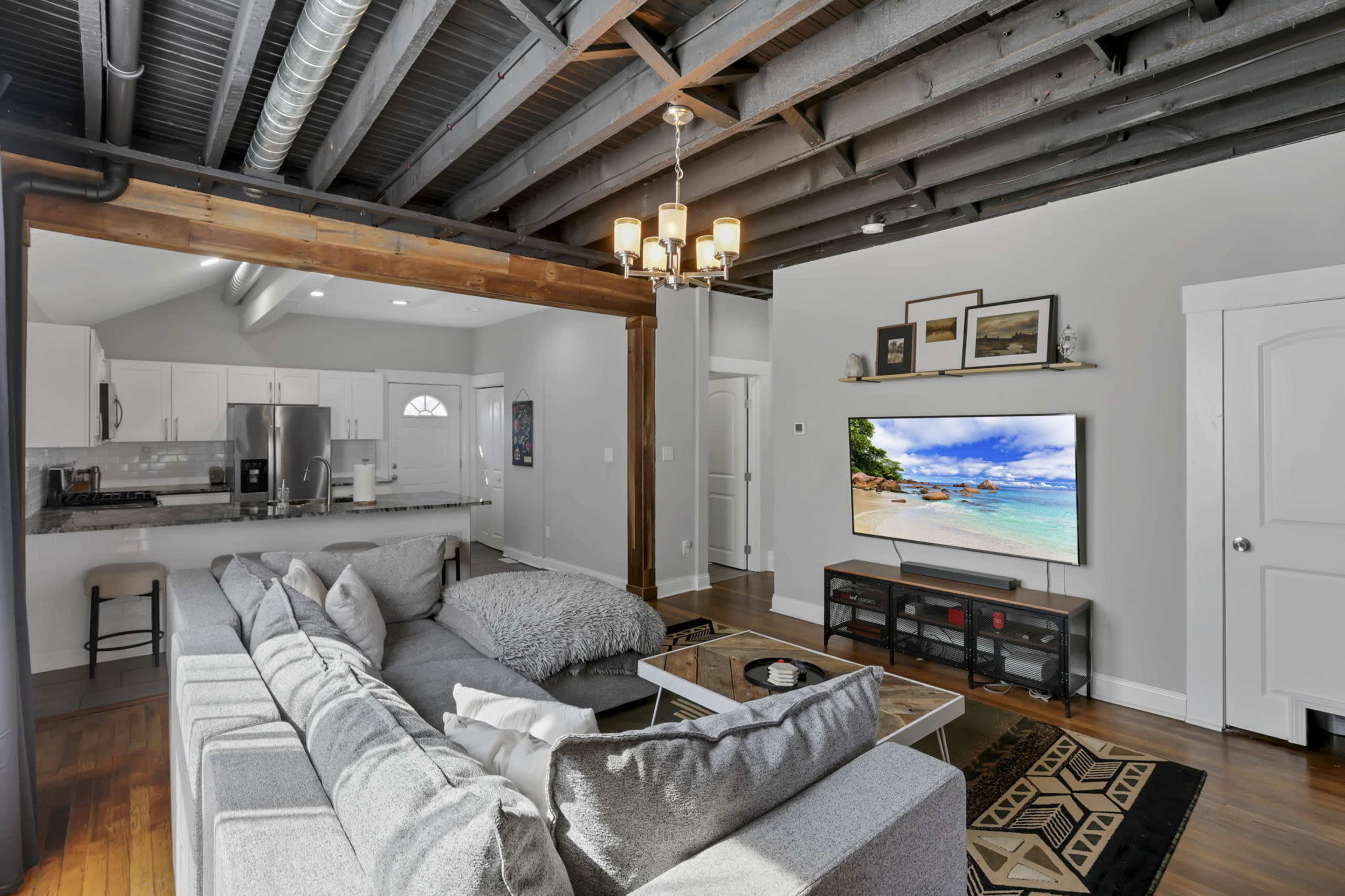 The image shows a modern living room featuring a gray sectional sofa, a coffee table, and a television displaying a beach scene, with exposed beams and a kitchen in the background.