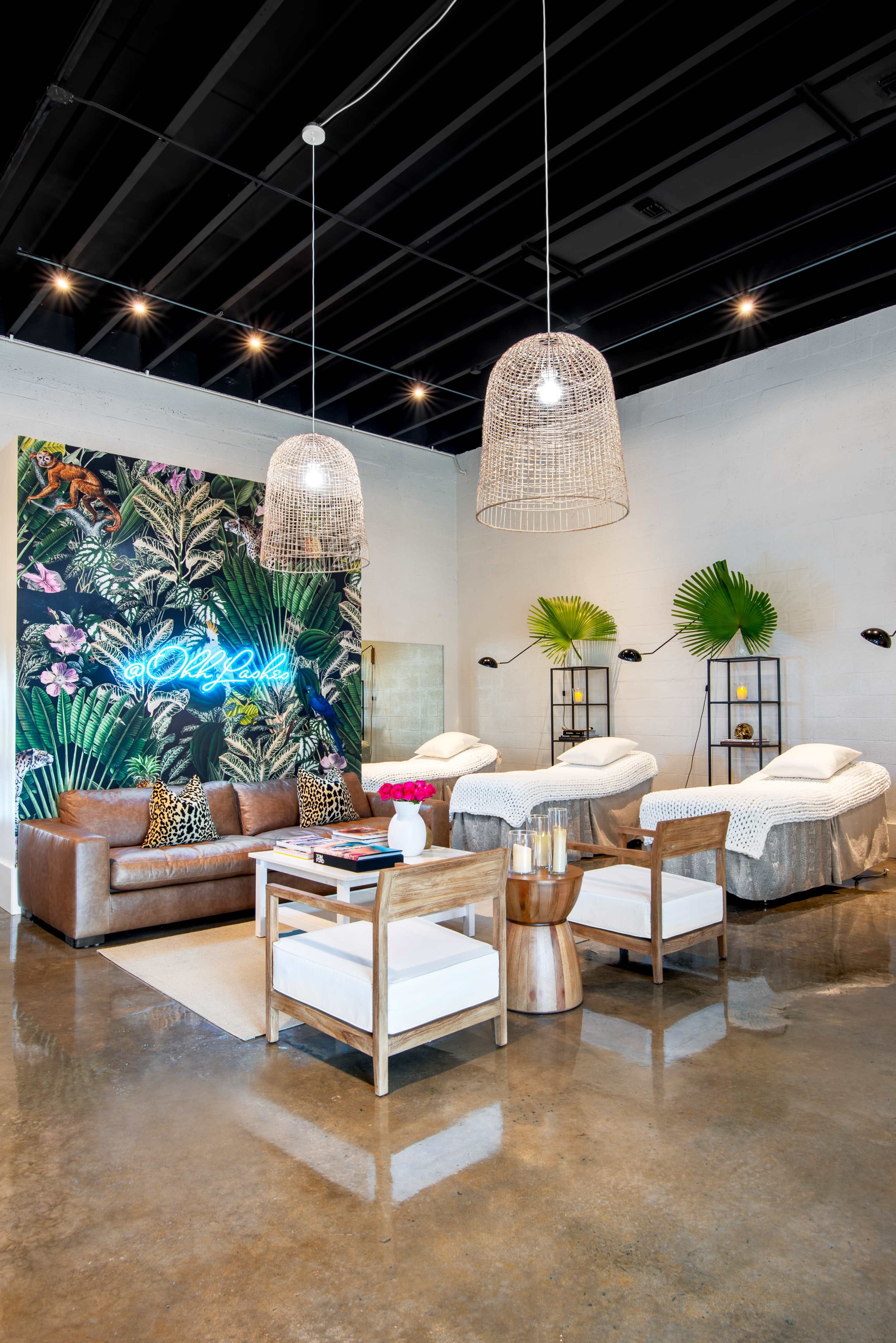 A modern relaxation area featuring a brown leather sofa, two treatment beds, and a vibrant tropical wall mural, illuminated by large hanging light fixtures.
