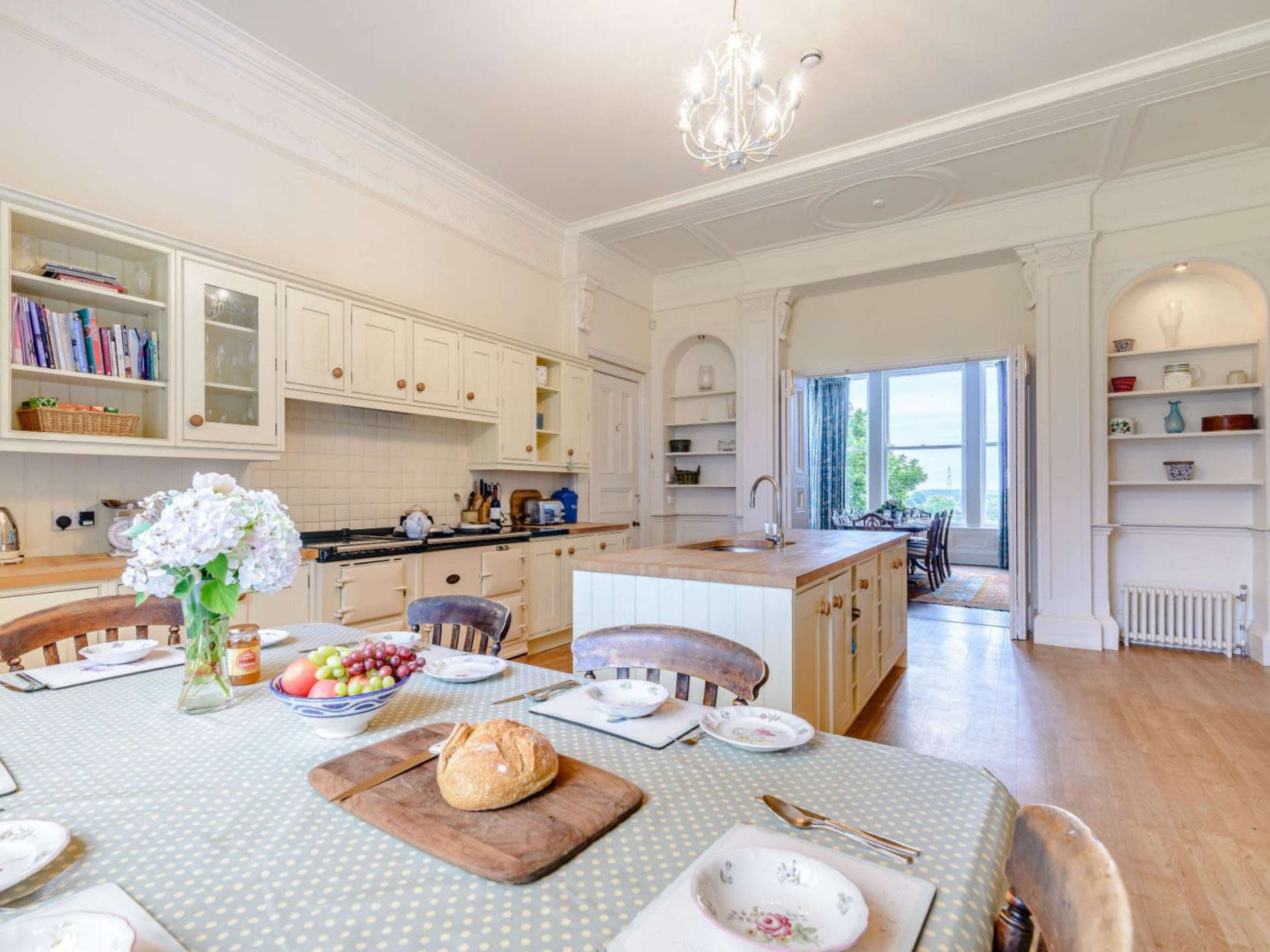 A spacious kitchen features a dining table set with plates and a loaf of bread, with an island and open shelves in the background.