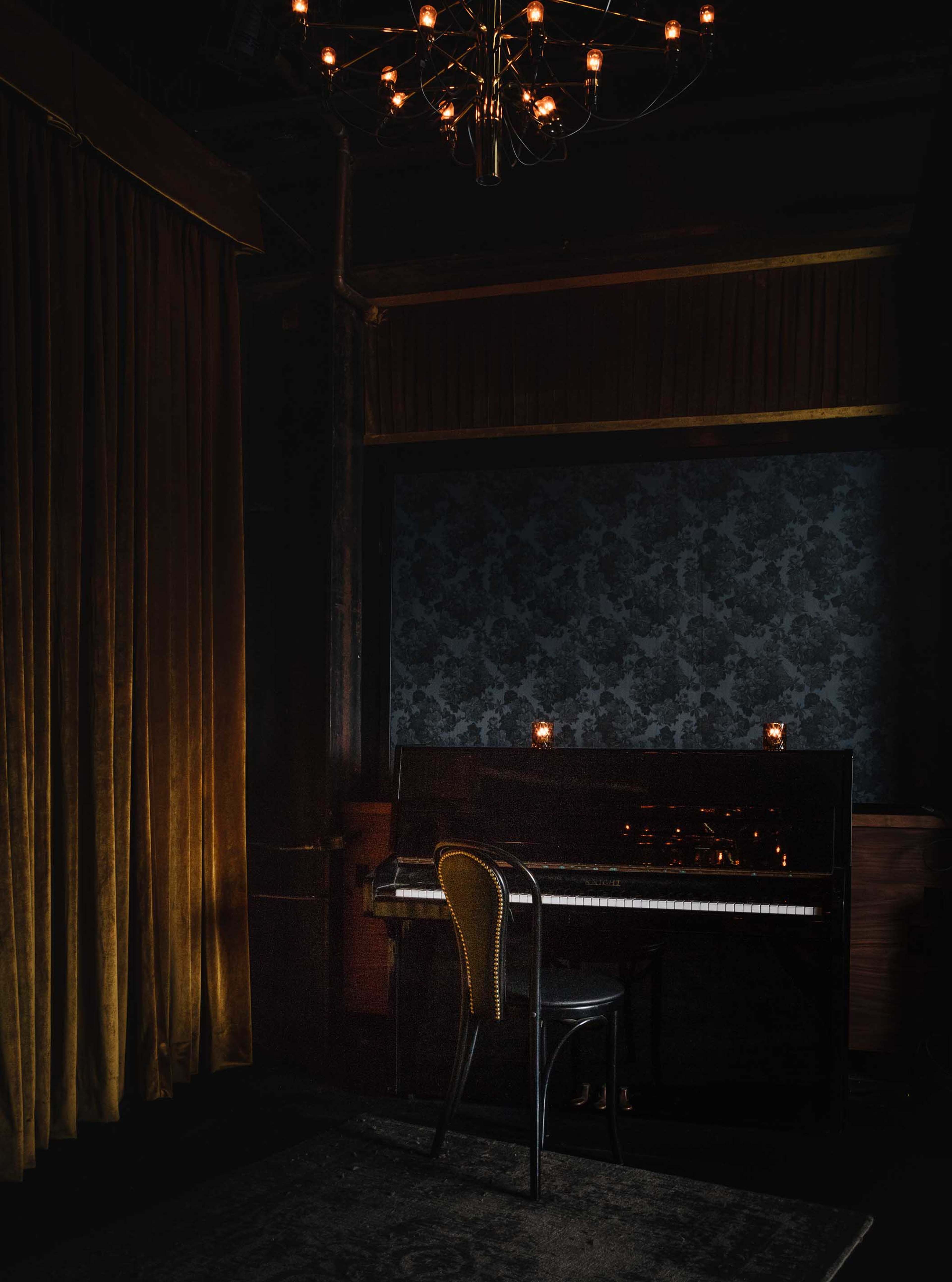 A grand piano sits in a dimly lit room, accompanied by a tall chandelier and a velvet curtain, with candles placed on the piano and wall.