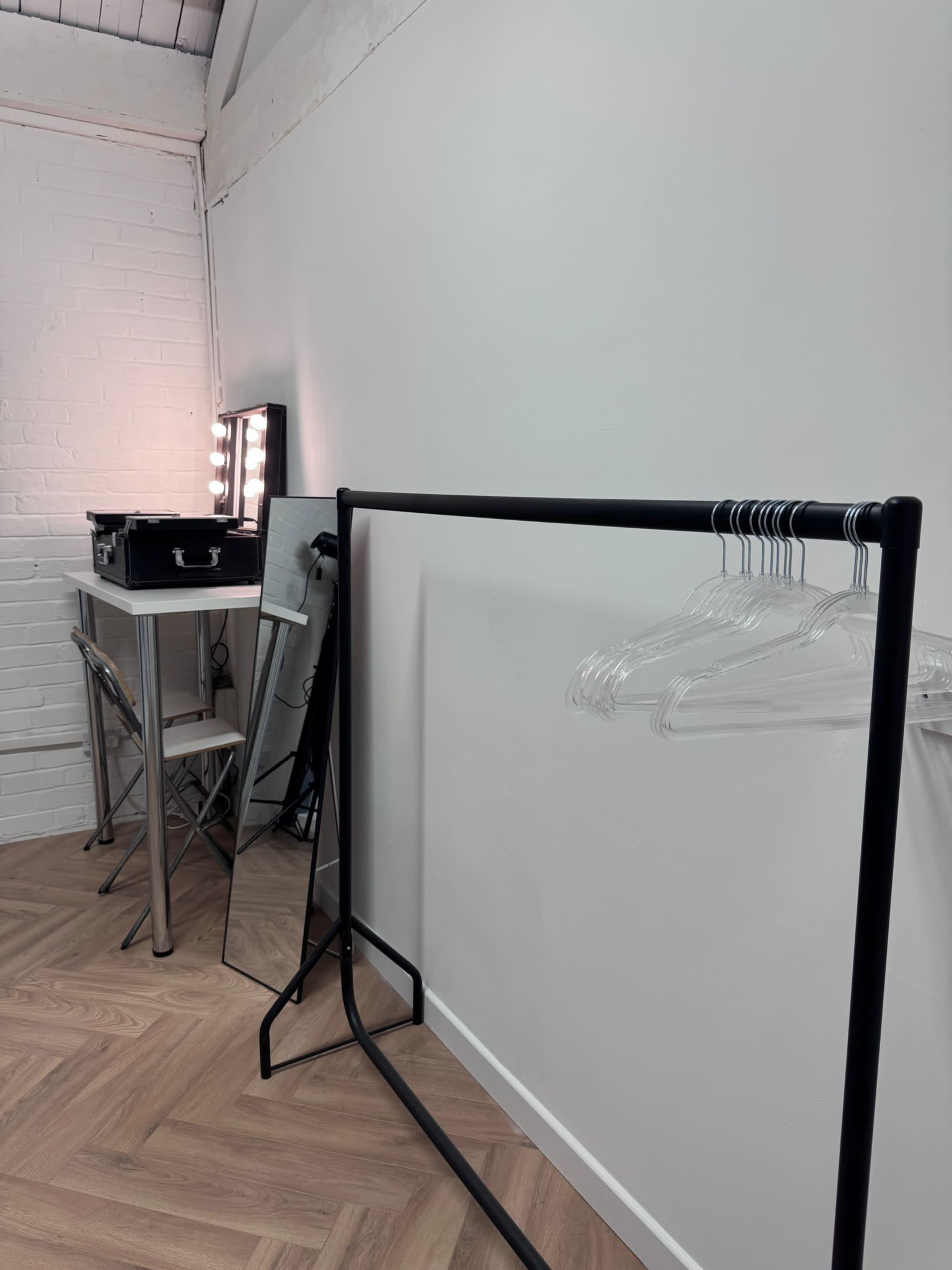 The image shows a minimalist room with a clothing rack, a table, and a mirror, all set against a white wall.