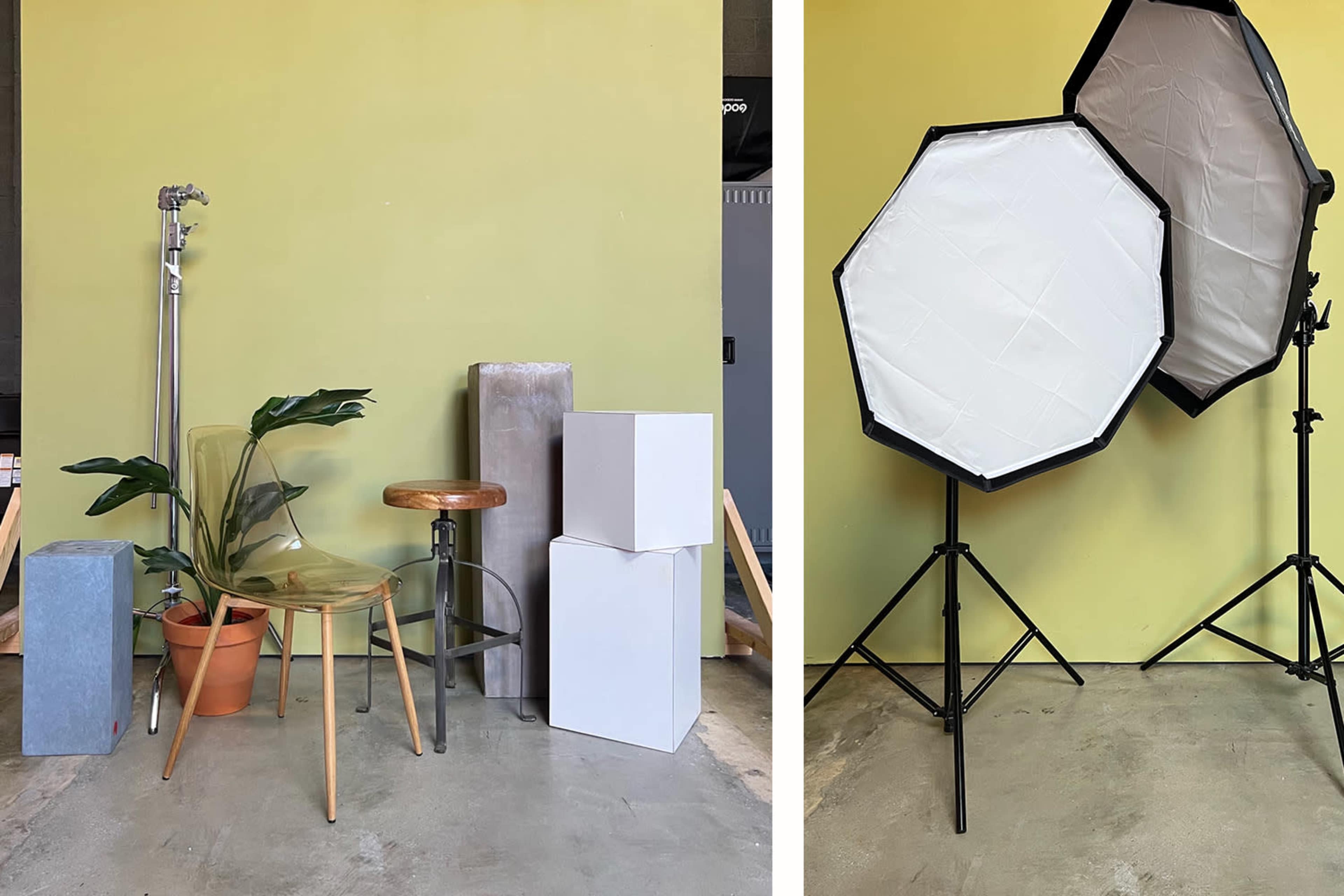 The scene features various studio props, including a clear chair, potted plant, cylindrical and rectangular boxes, and two softboxes on stands against a green wall.