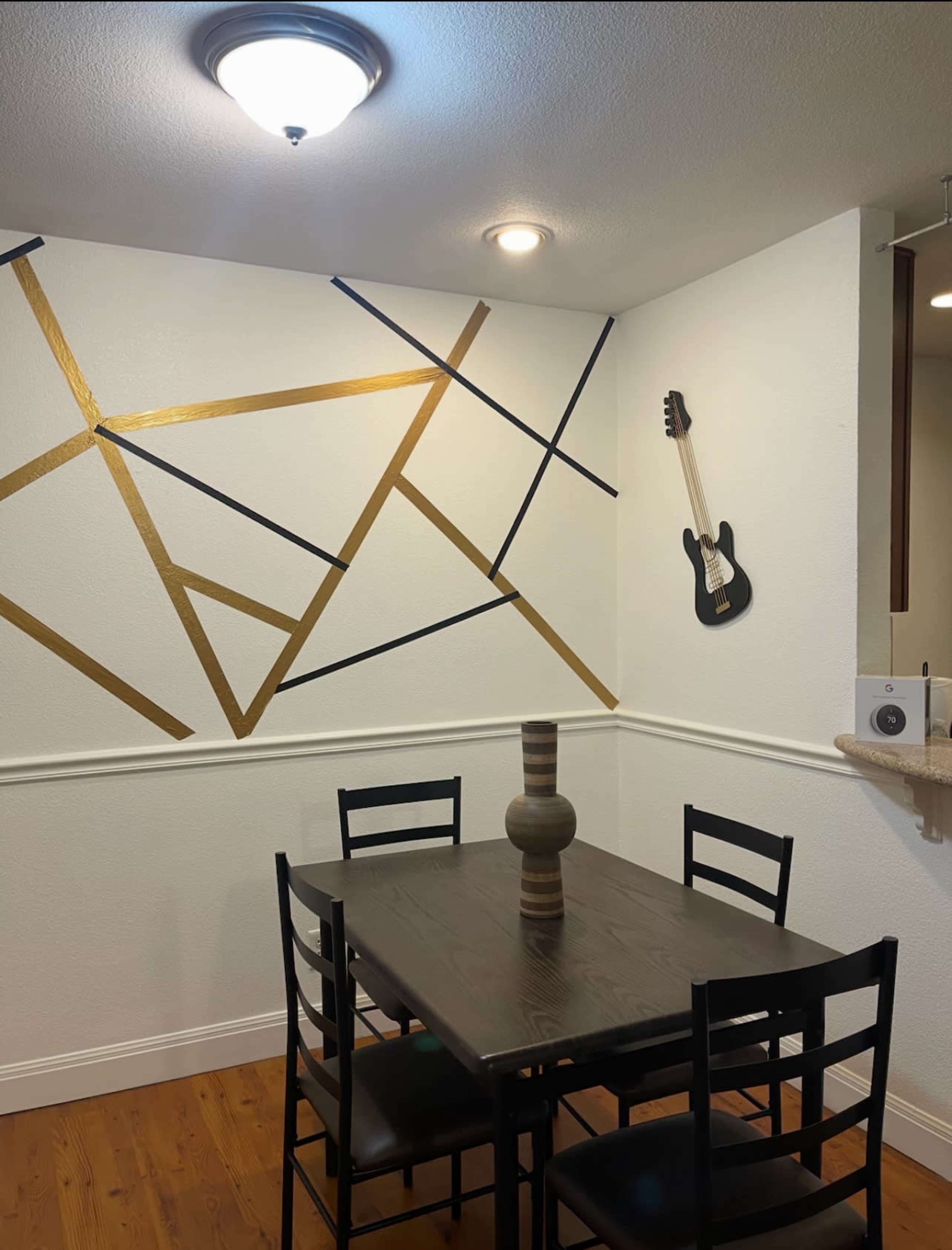 The image shows a dining area with a black table and chairs, a decorative wall featuring abstract patterns in gold and black, and a guitar mounted on the wall.
