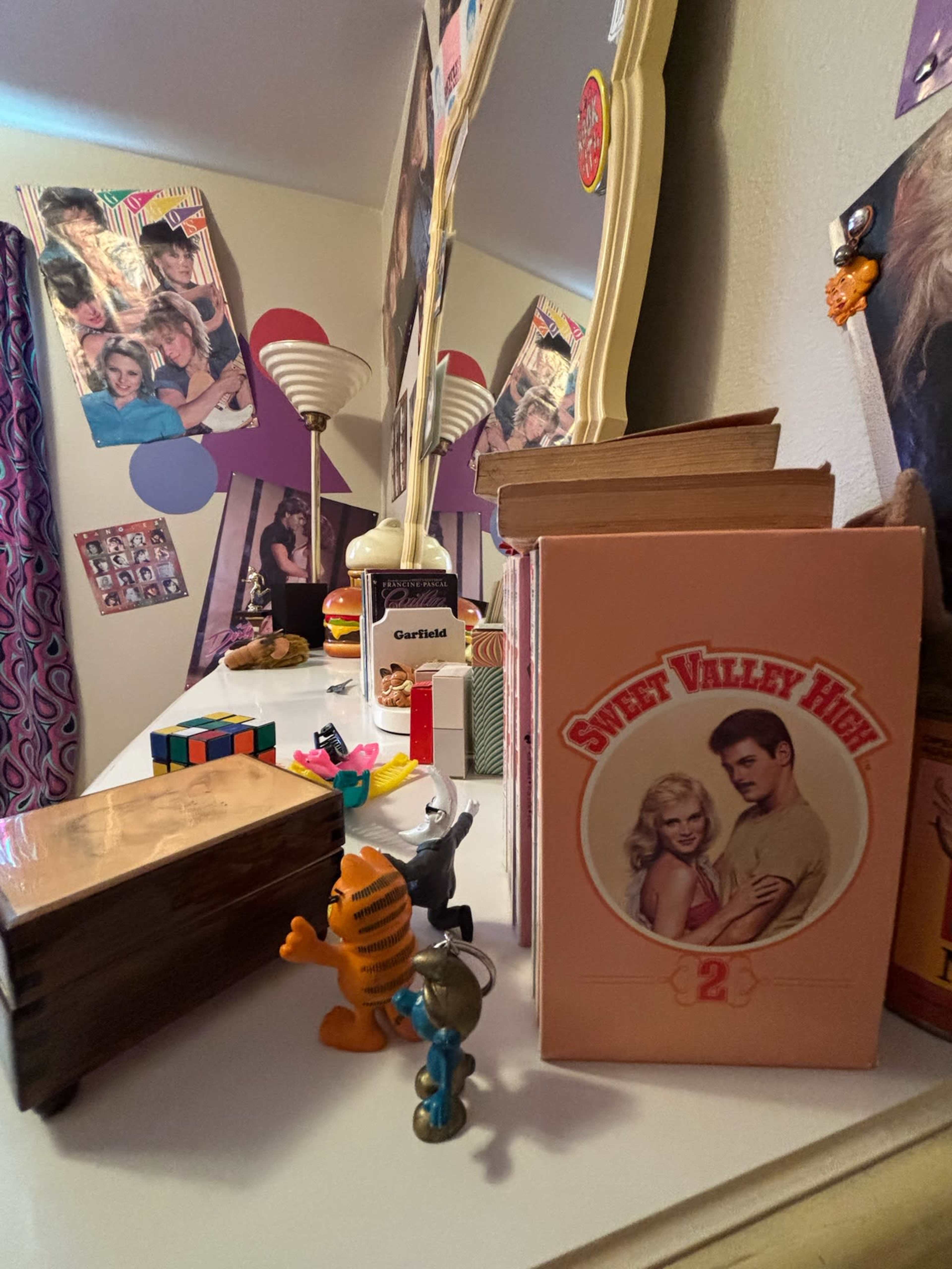 A cluttered dresser displays vintage books, toys, and decorations, with a mirror reflecting posters on the wall.