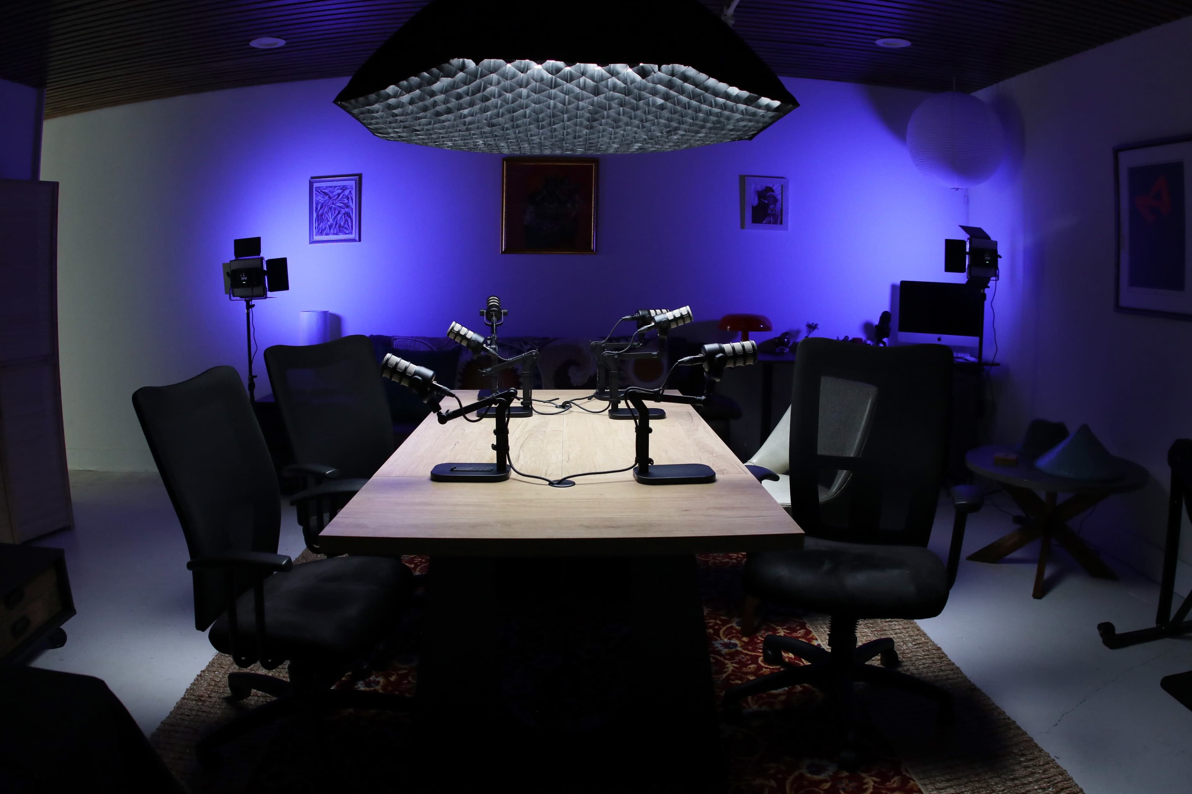 A podcast recording studio features a wooden table with two microphones and five black chairs, illuminated by purple lighting.