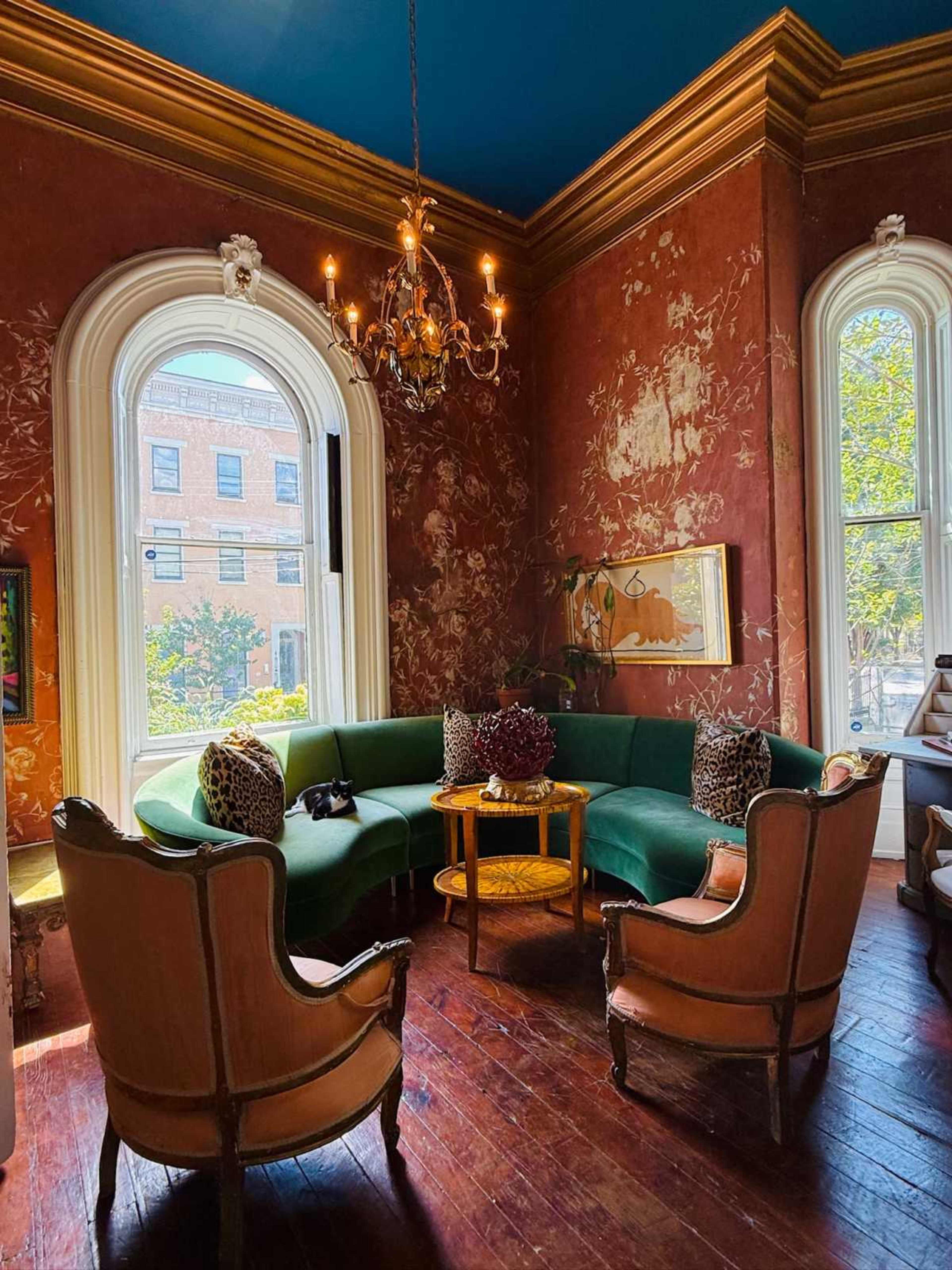 The image shows a cozy corner of a room featuring a curved green sofa, two orange armchairs, a small round table, and large windows with natural light.