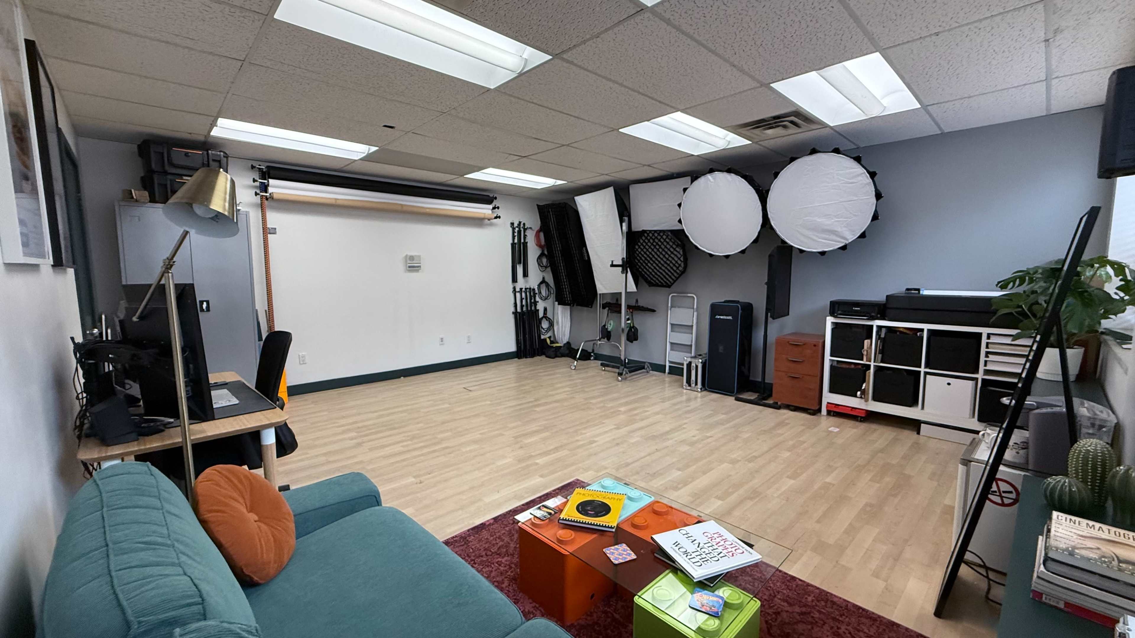 The image shows a spacious studio with wooden flooring, a green couch, a desk with a computer, and various photography equipment arranged along the walls.