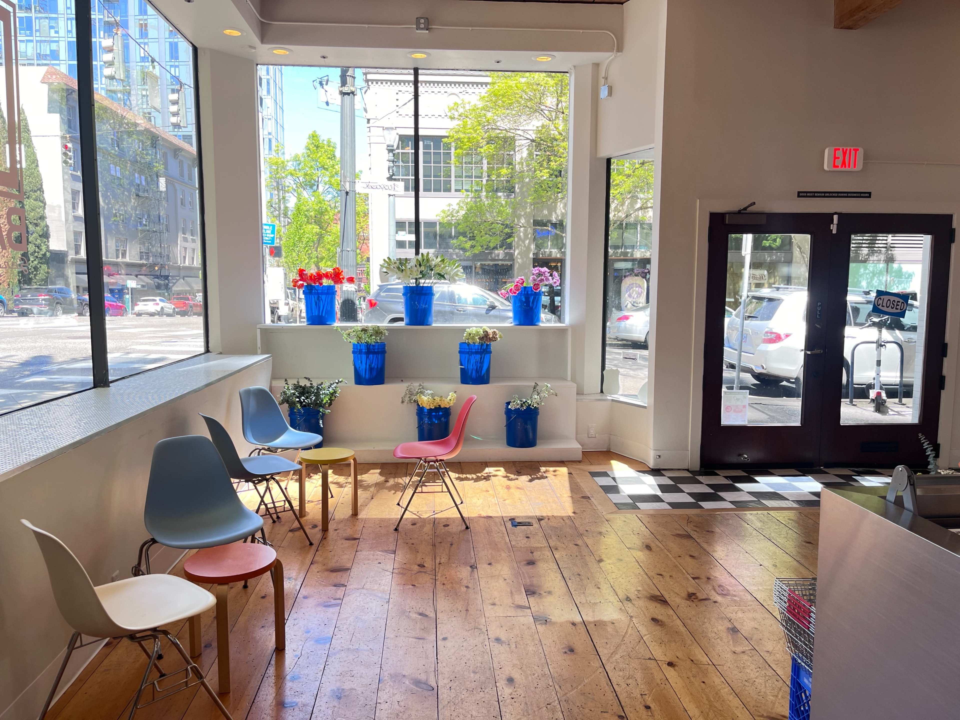 A bright café interior features colorful chairs and potted plants arranged by large windows, with a view of a bustling street outside.