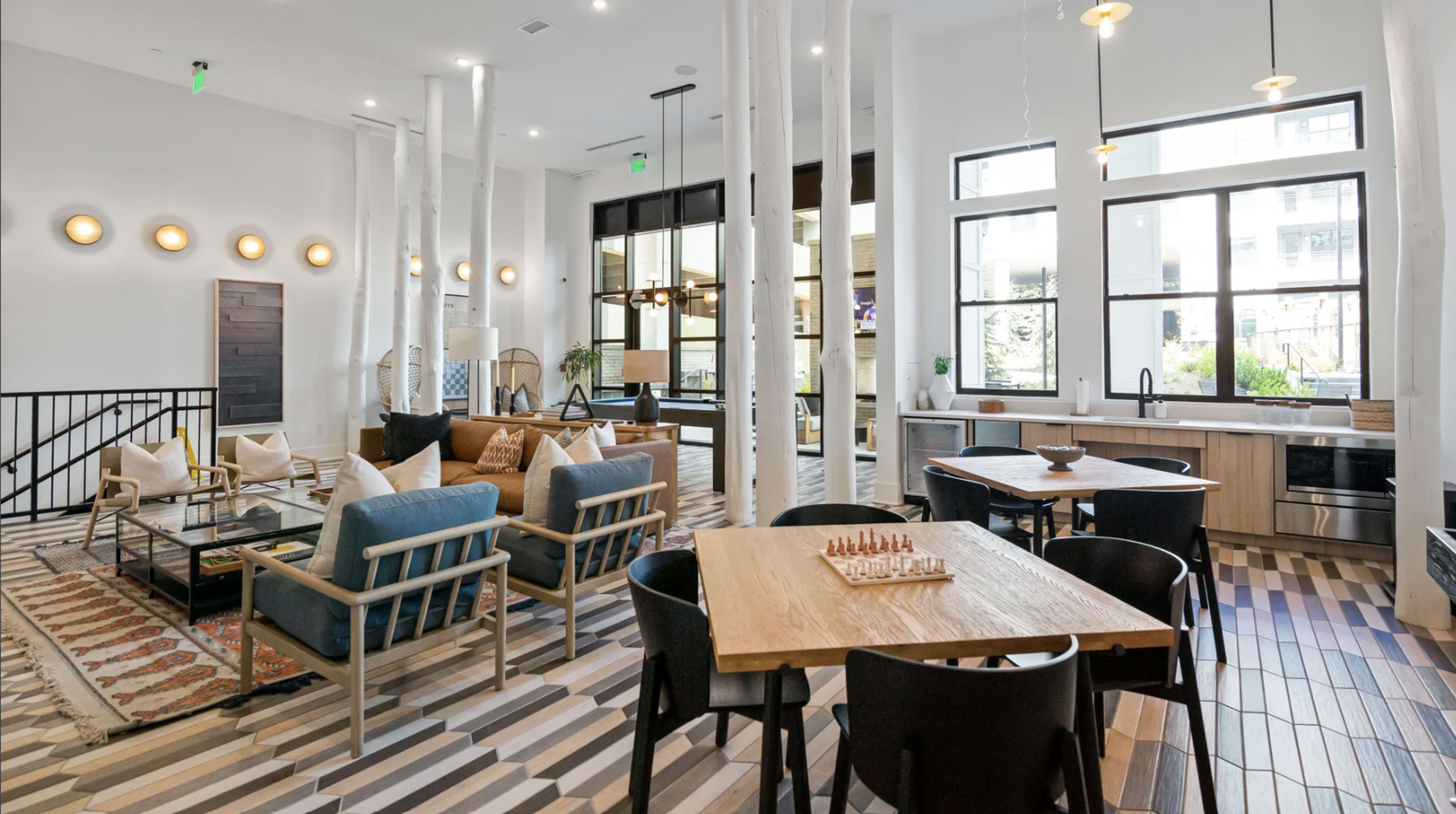 The image depicts a modern interior space featuring a lounge area with couches, a dining table, and a kitchen in an open layout with large windows.