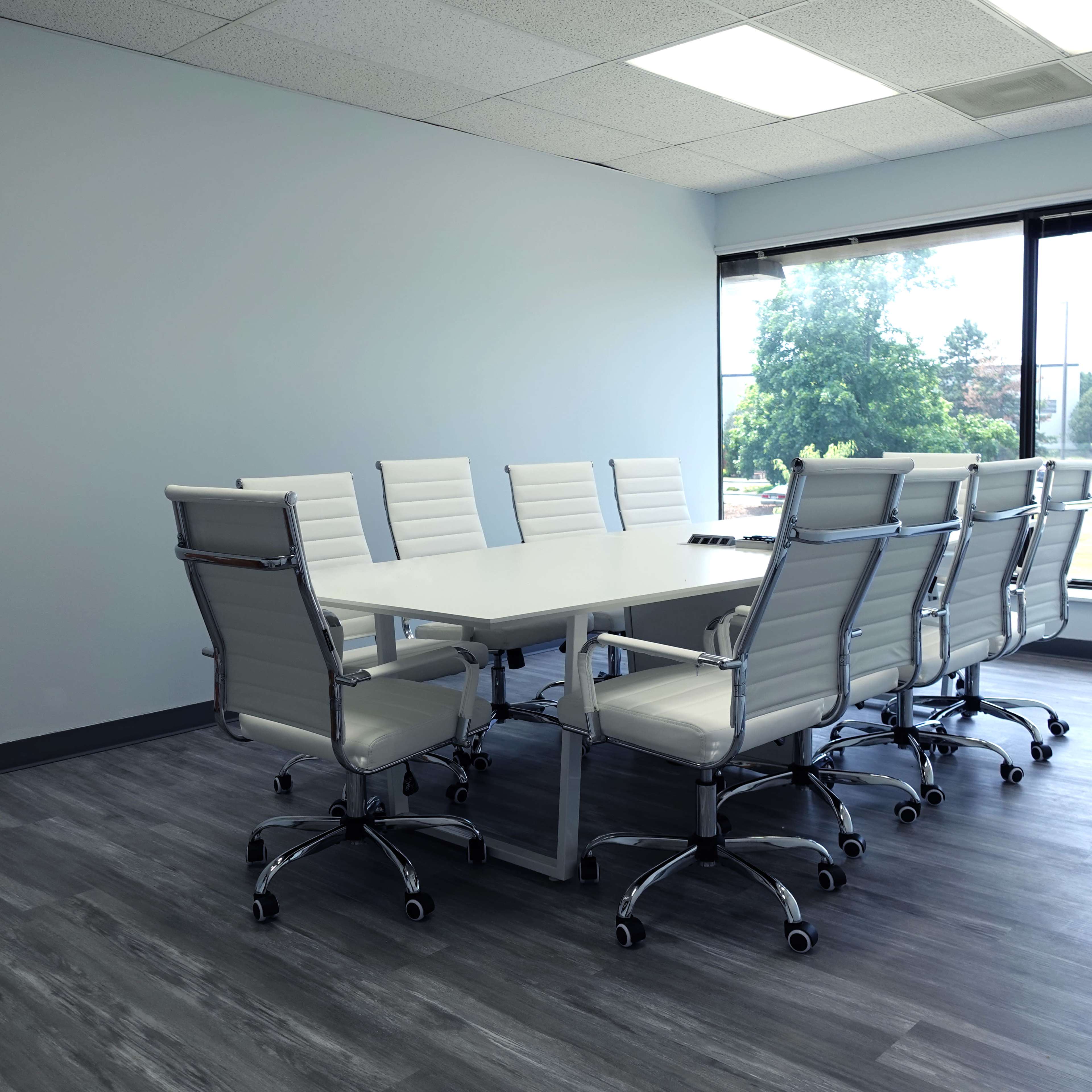 The image shows a modern conference room with a long white table surrounded by eight white rolling chairs and large windows allowing natural light.