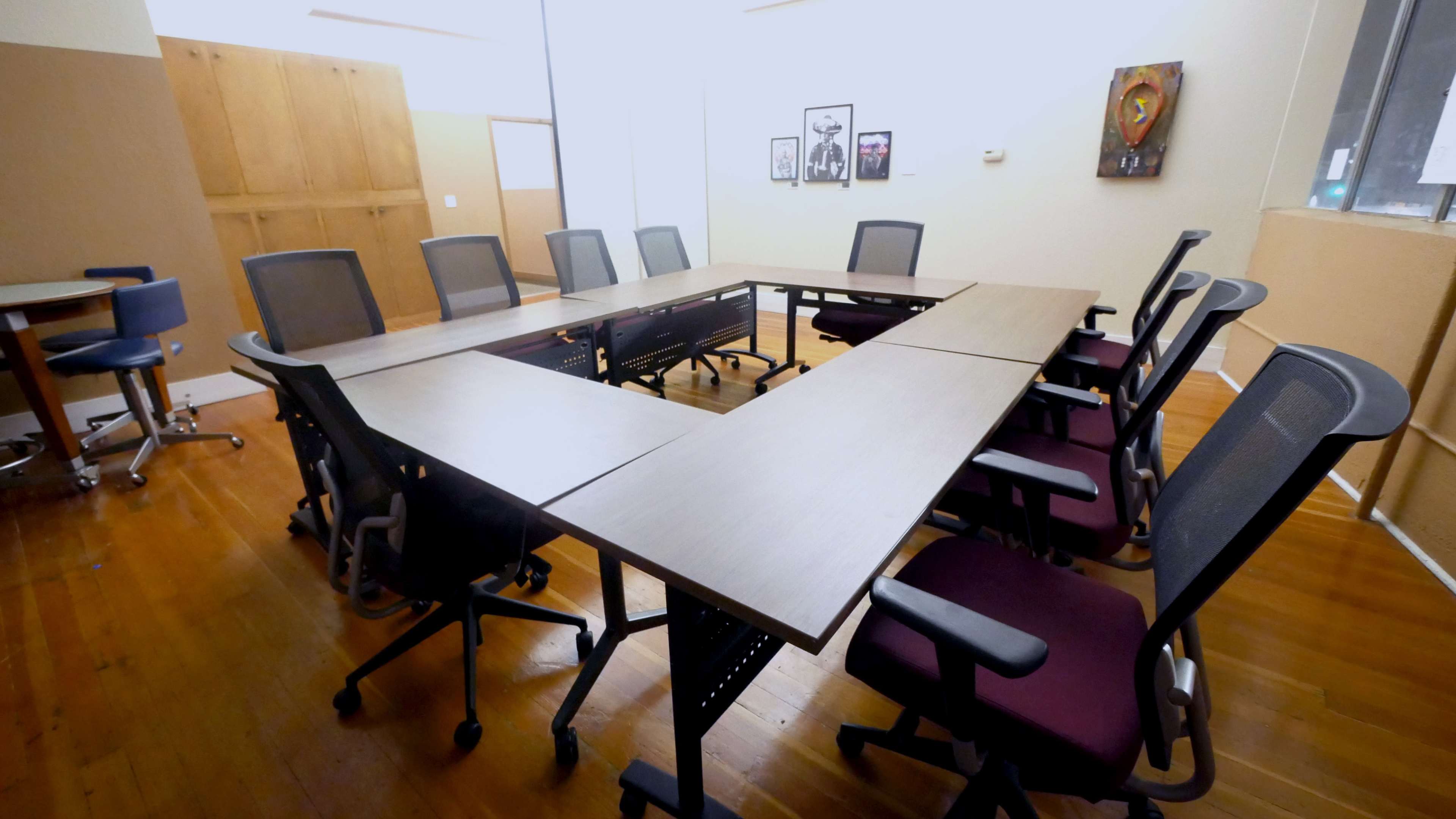 A conference room features a U-shaped arrangement of tables surrounded by rolling chairs, with wooden flooring and minimal decorations on the walls.