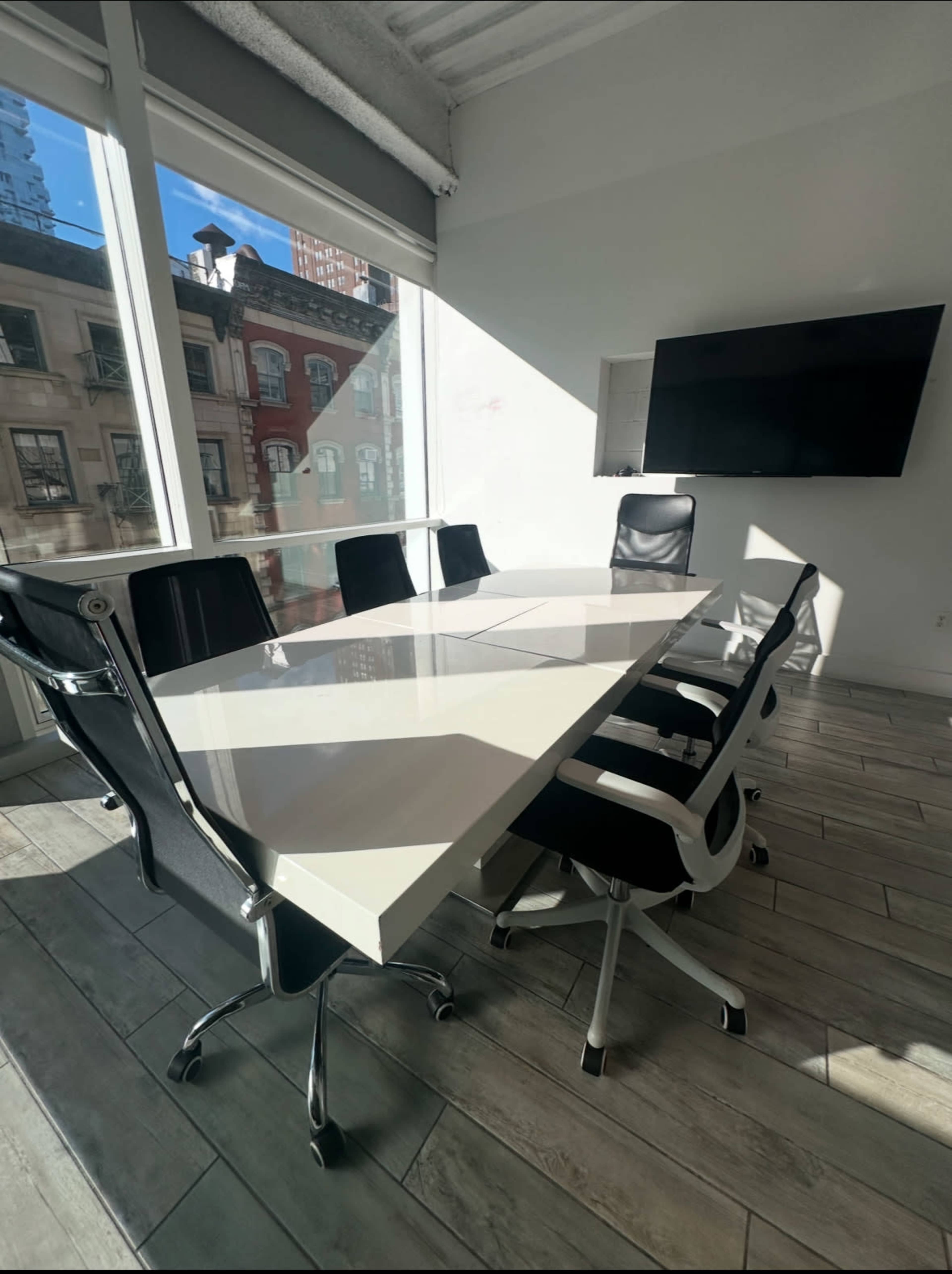 The image shows a modern conference room with a white rectangular table and several black and white ergonomic chairs, illuminated by natural light from large windows.