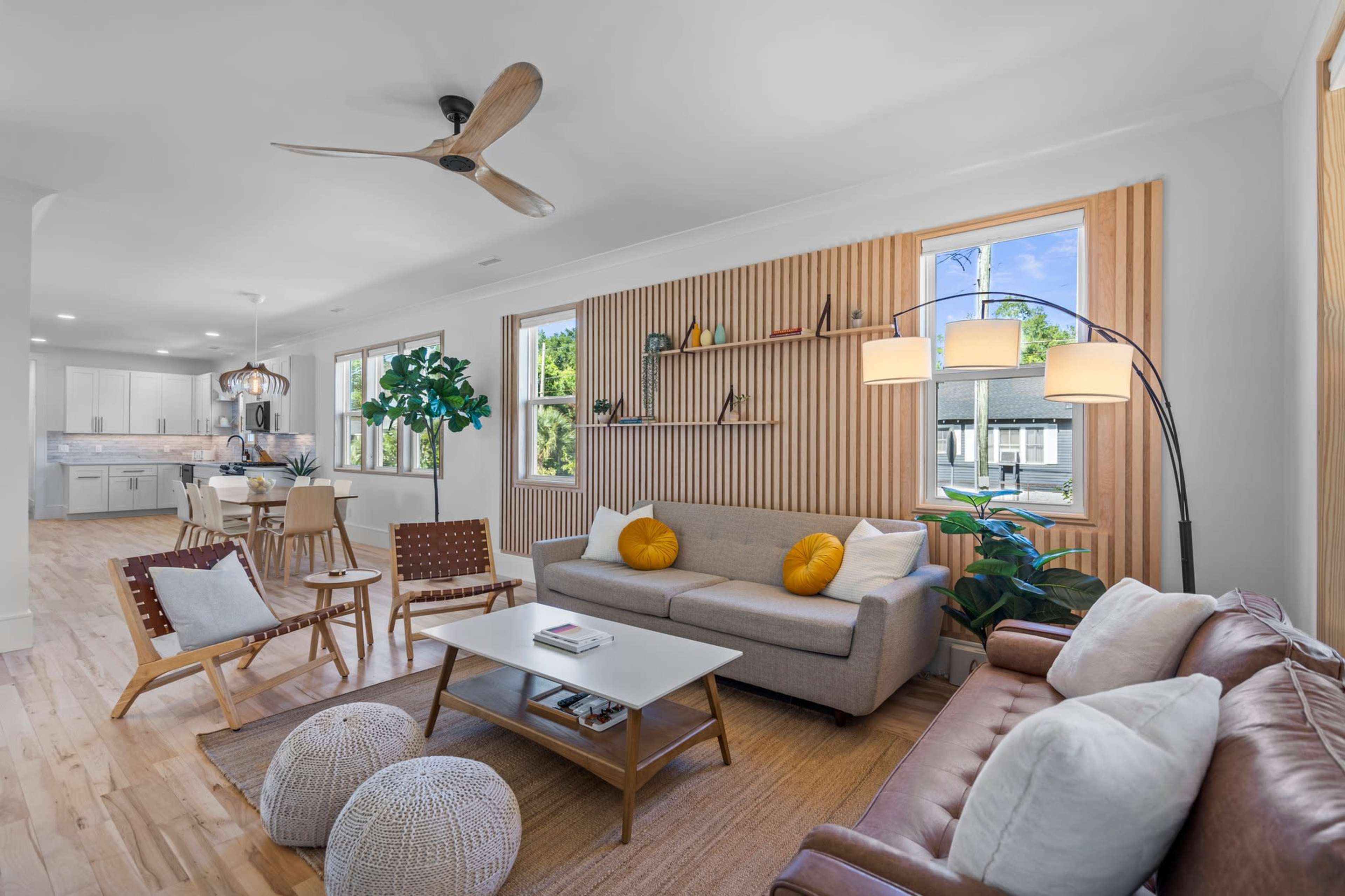 A modern living room with a gray sofa, a brown leather couch, wooden shelving, and large windows allowing natural light to enter.