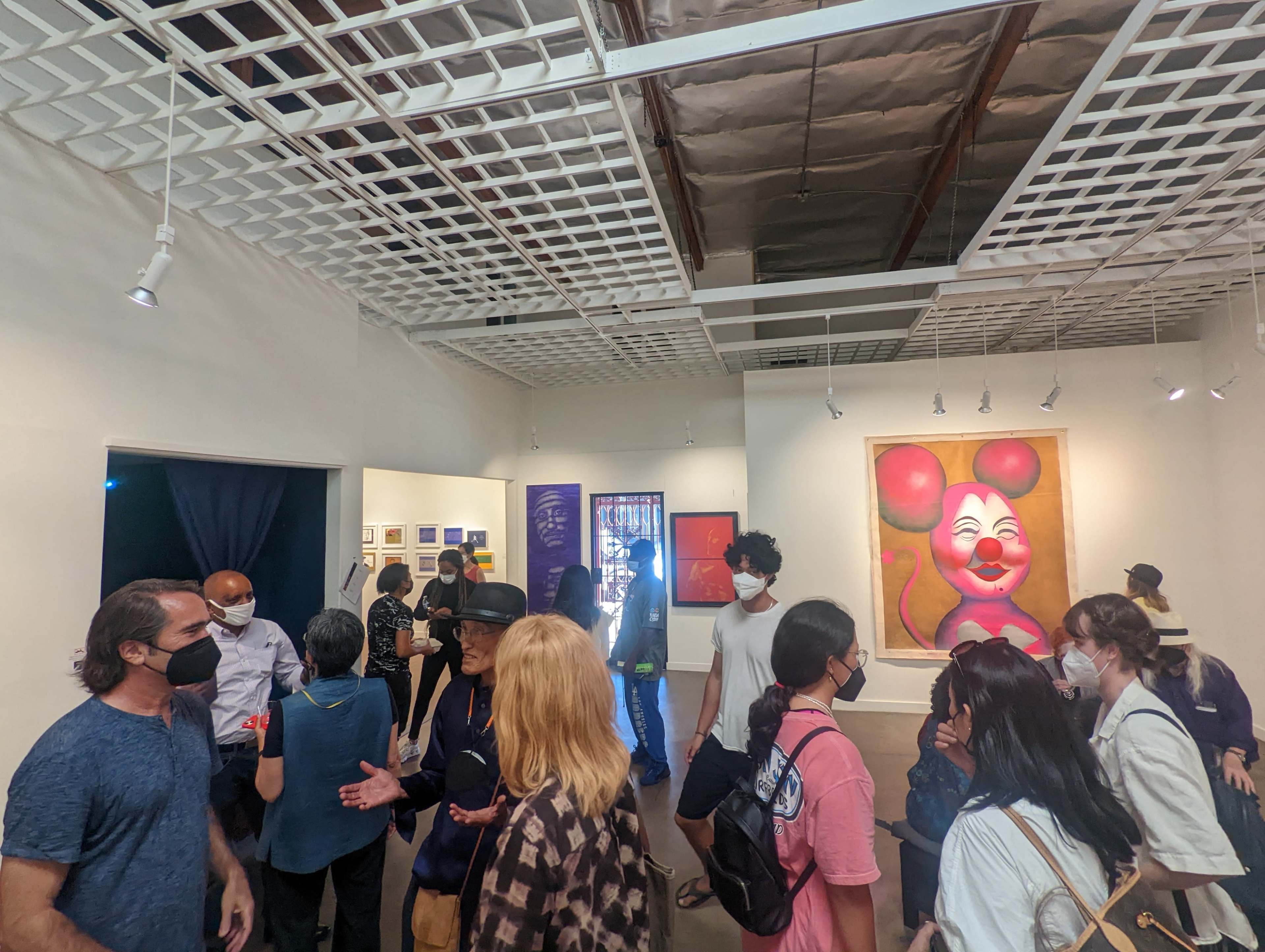 A group of people in masks engages in conversation at an art gallery featuring colorful artwork on the walls.