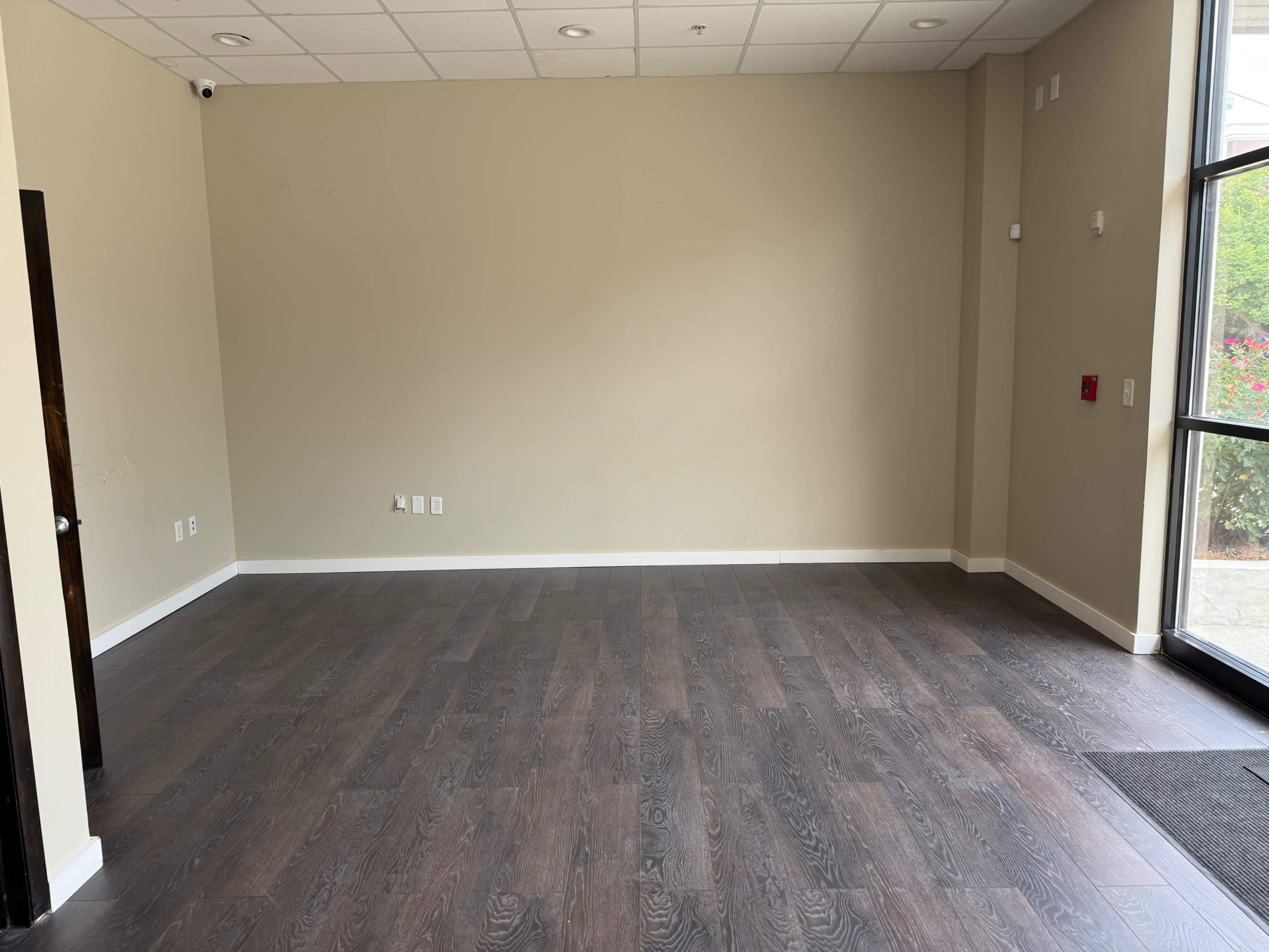 The image shows an empty room with brown wooden flooring and light beige walls, featuring a large window.