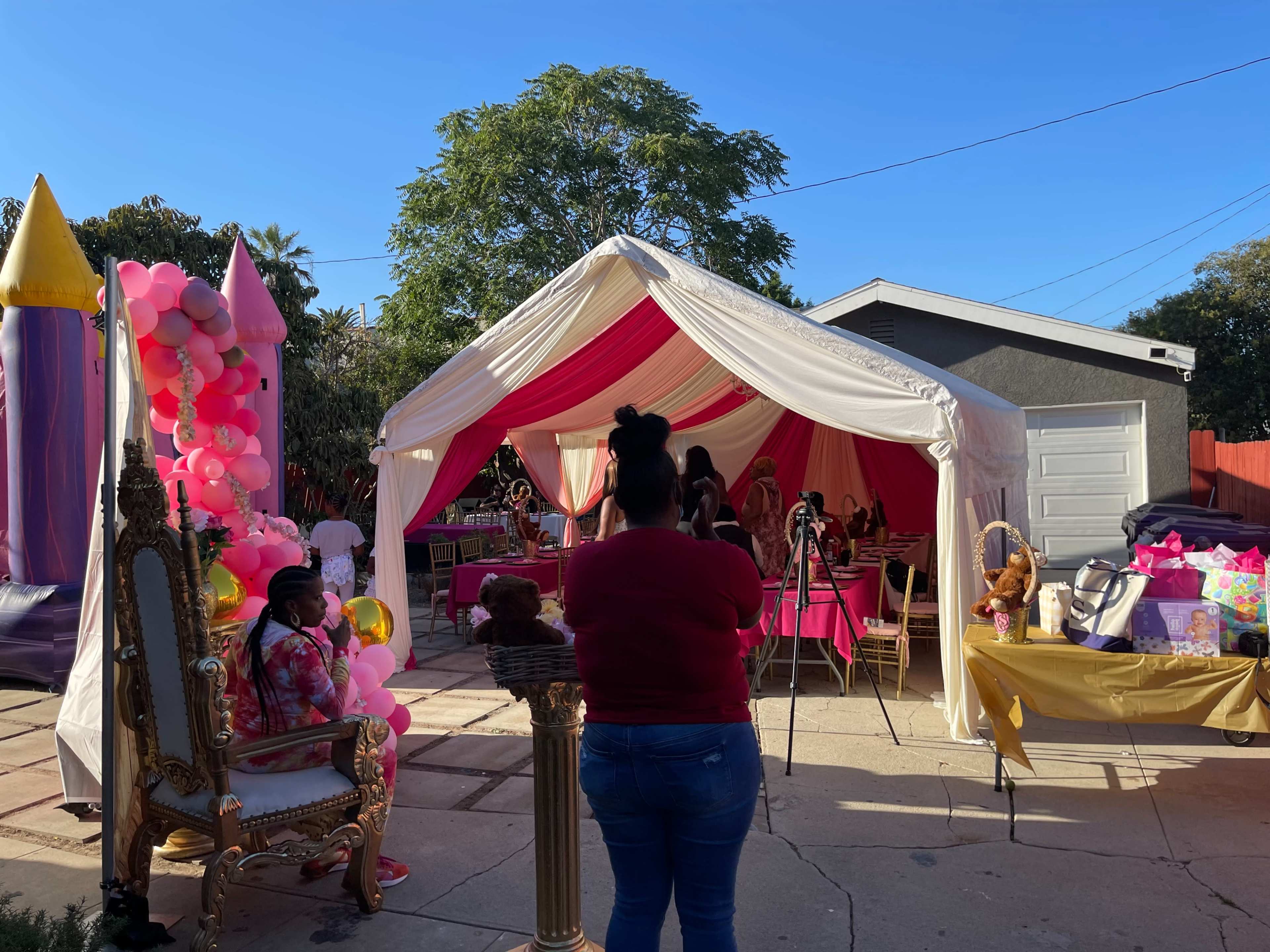 A tent with pink and white decorations hosts a celebration filled with balloons, gifts, and chairs in a backyard setting.