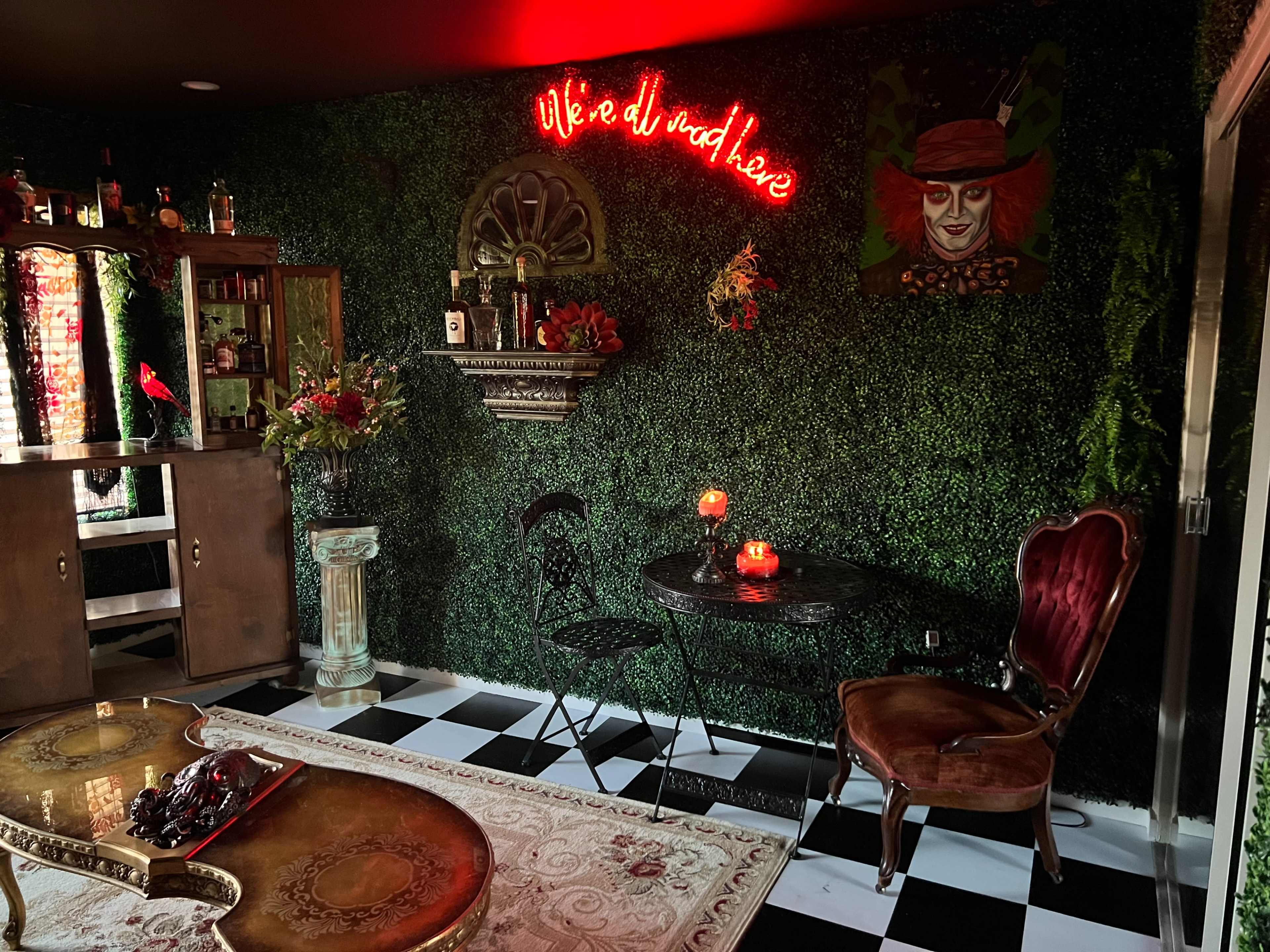 The interior features a dark green hedged wall, a small table with candles, a vintage chair, and a bar area decorated with various bottles and a colorful painting.