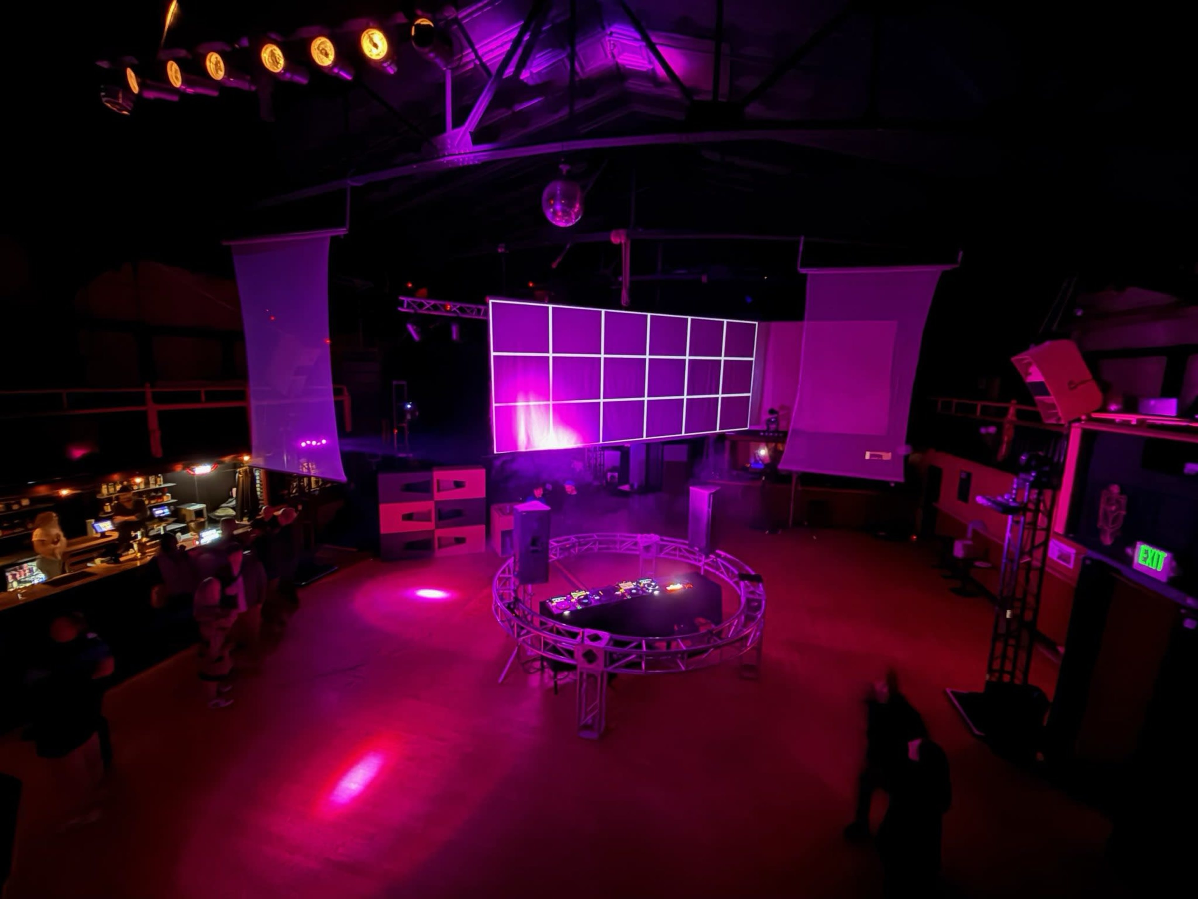 The image shows a dimly lit event space featuring a large video screen displaying a grid pattern, with colorful lighting and smoke effects enhancing the atmosphere.
