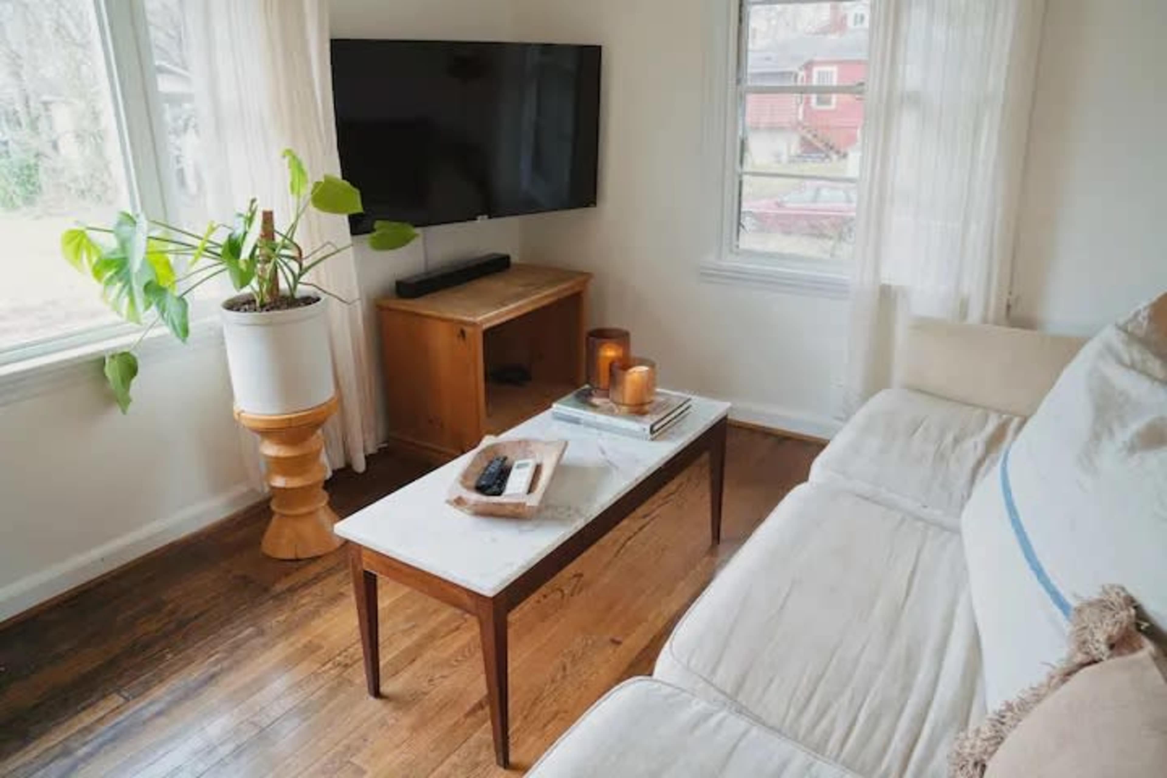A living room features a white sofa, a wooden coffee table, a TV mounted on the wall, and a potted plant beside a window.