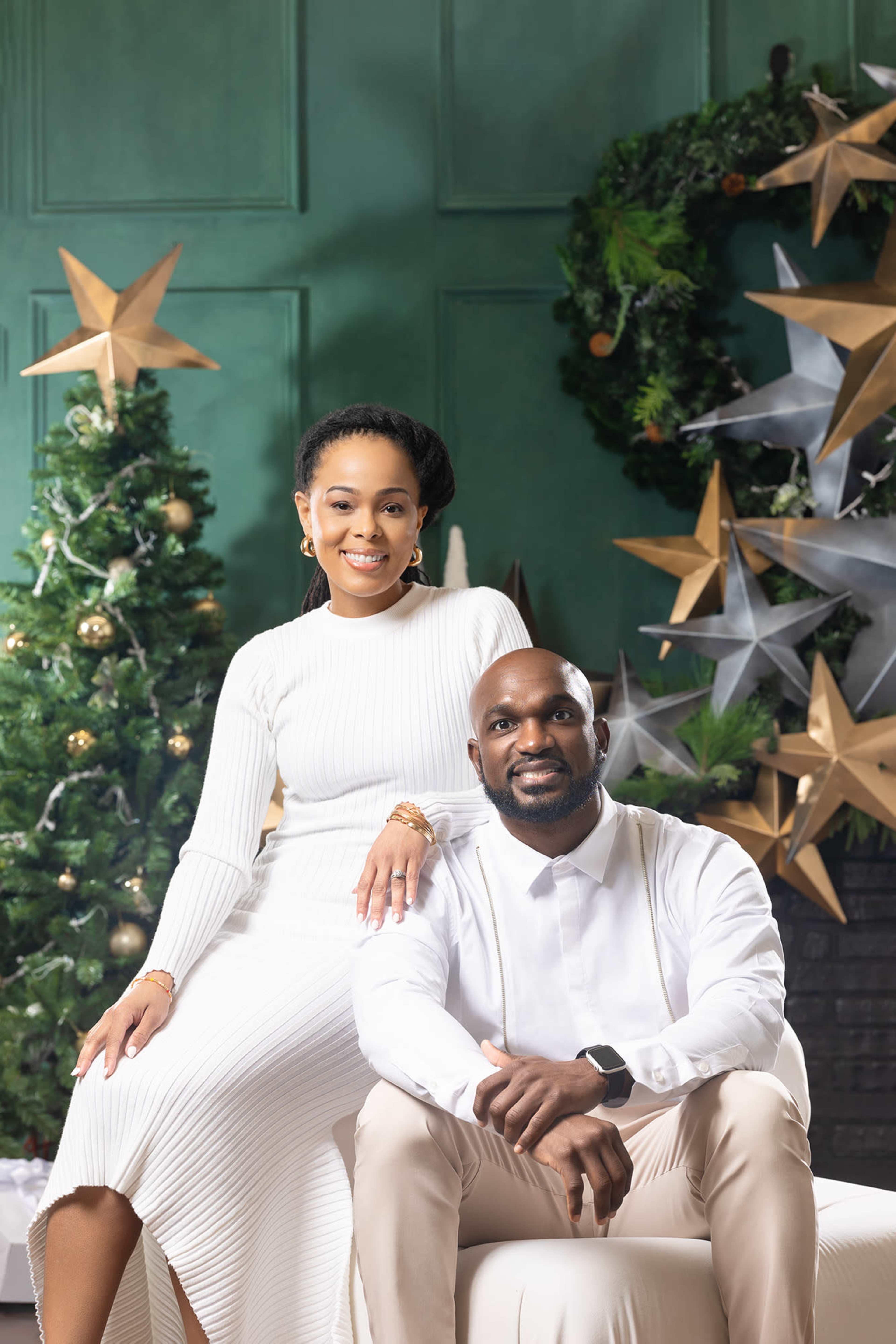 A man sits on a sofa next to a woman who is standing beside him, both are dressed in formal attire, with a decorated Christmas tree and festive ornaments in the background.