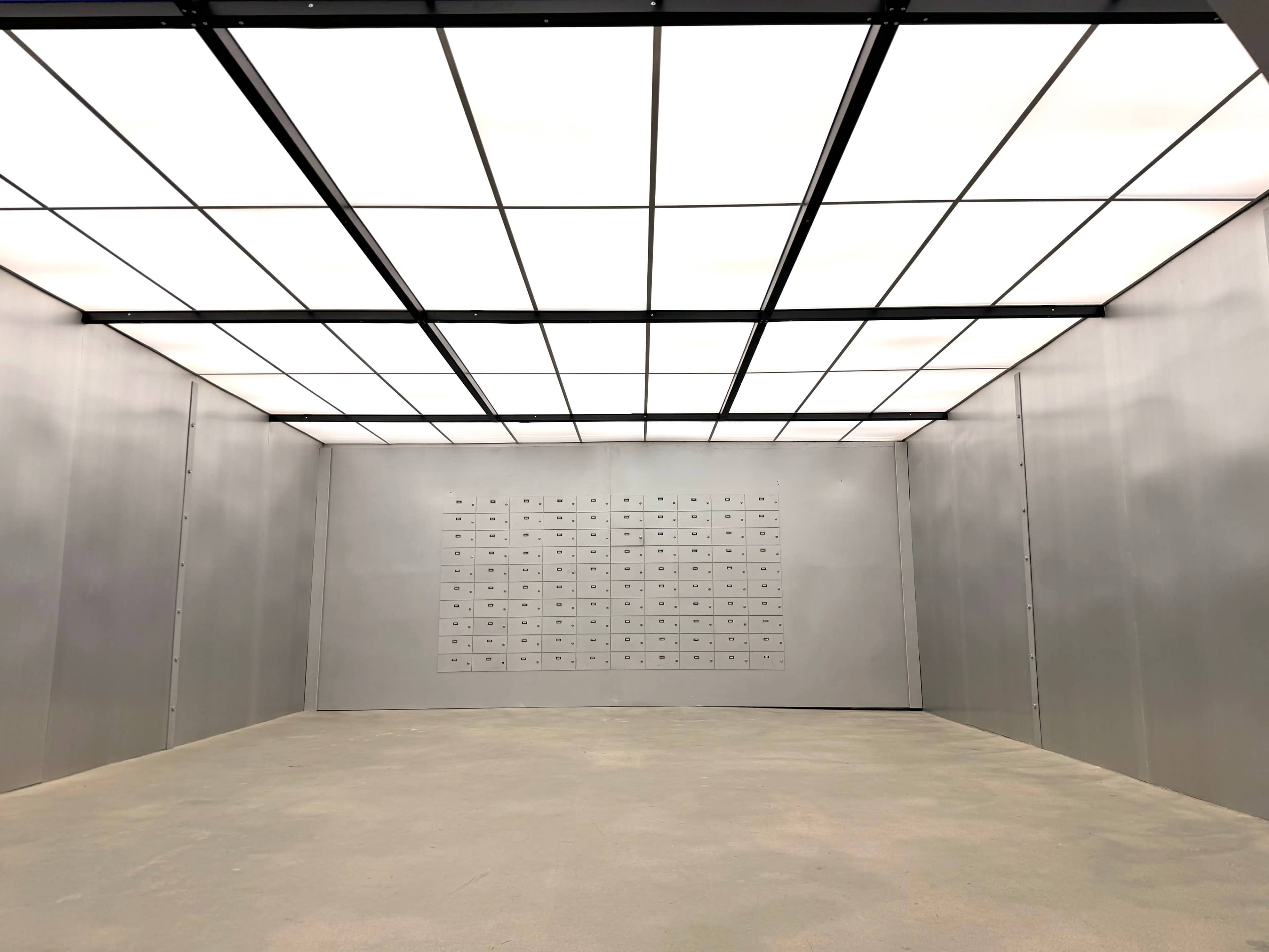 The image shows a large, empty room with gray walls and a grid of small, rectangular boxes mounted on one wall, illuminated by bright overhead lighting.