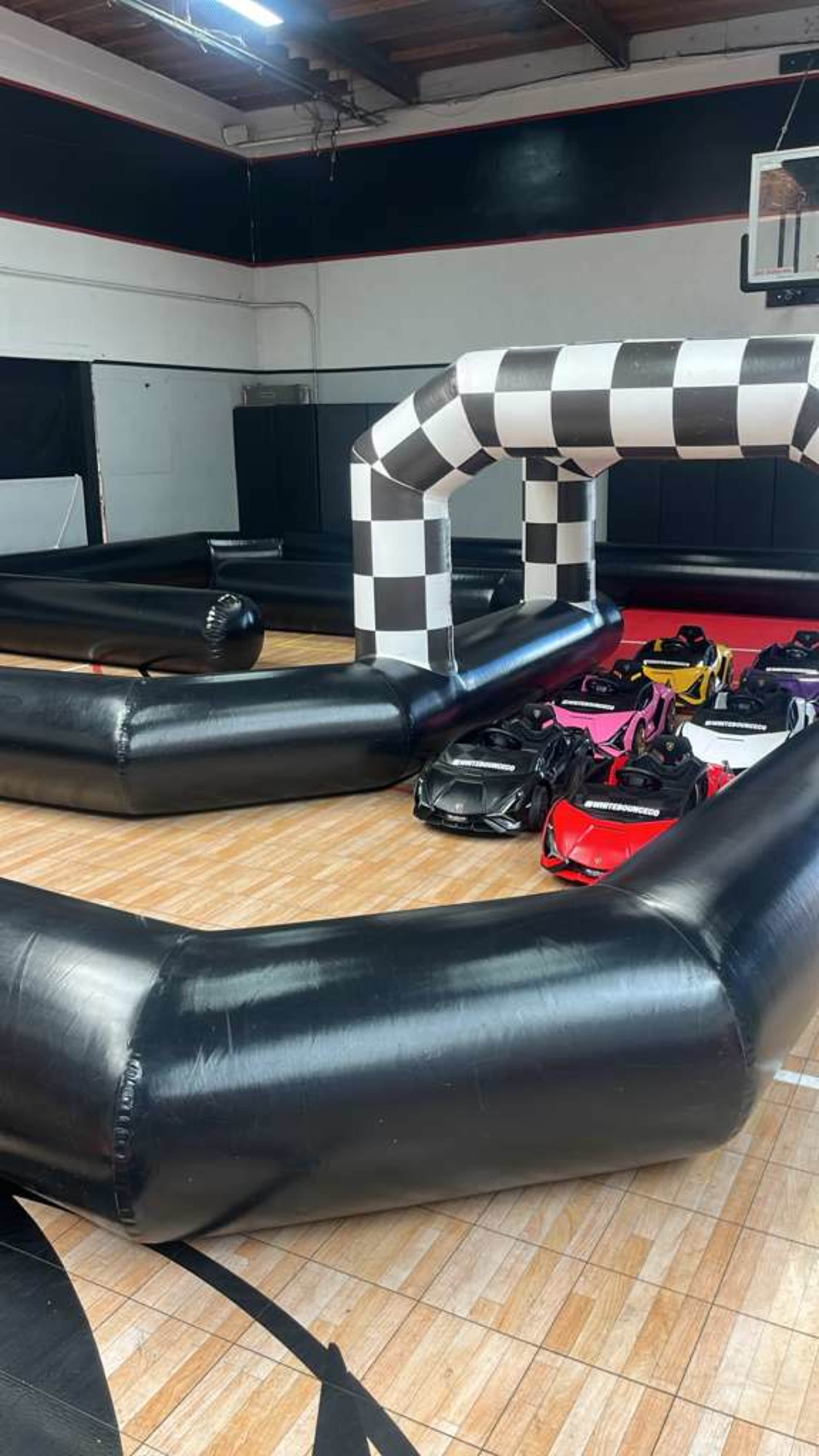 The image shows a black and white checkered inflatable race track set up on a wooden gym floor, with bags of equipment placed beside it.