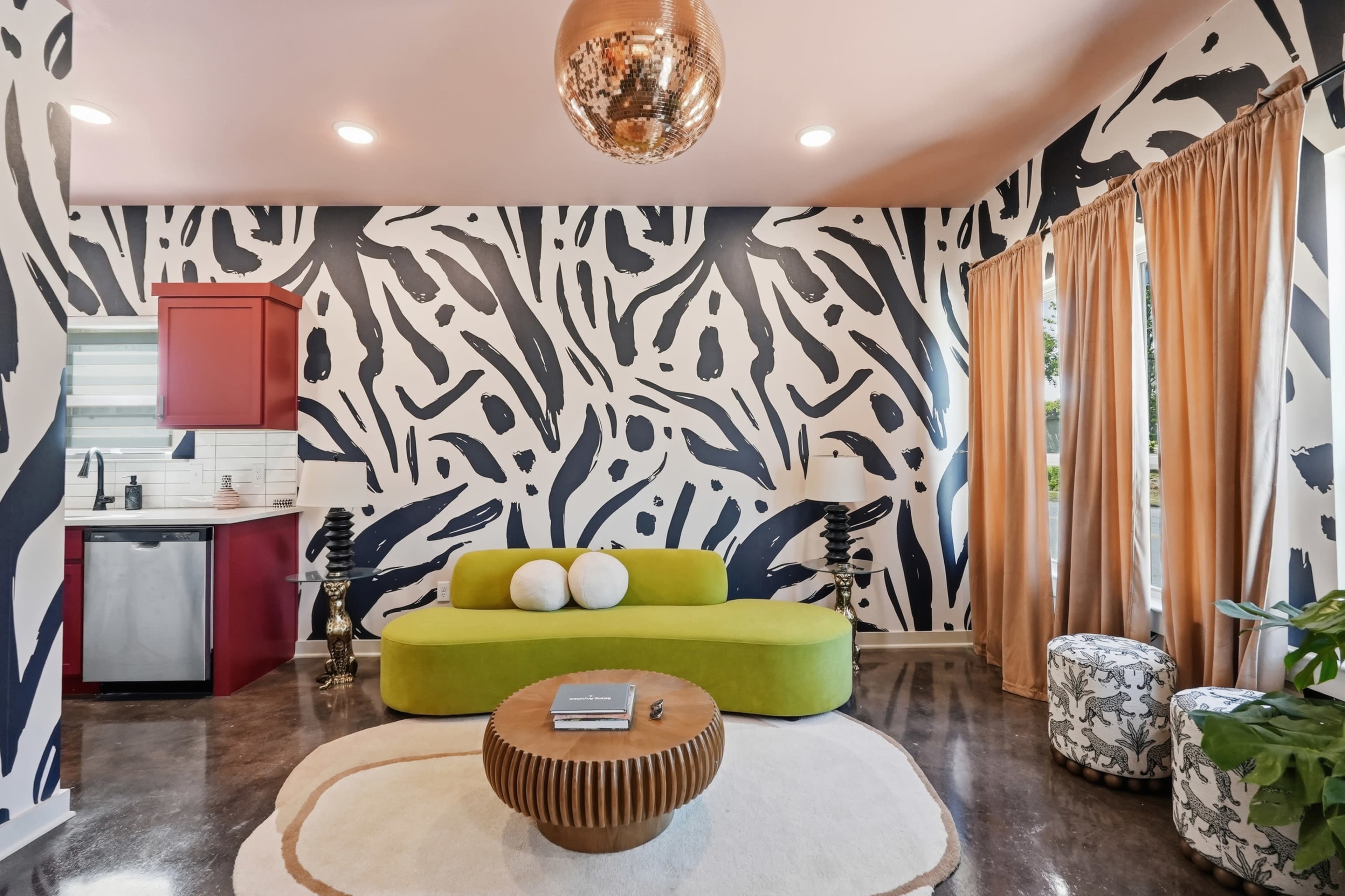 The image shows a modern living room featuring a green sofa, a round coffee table, and bold patterned wallpaper, with curtains and decorative stools adding to the decor.