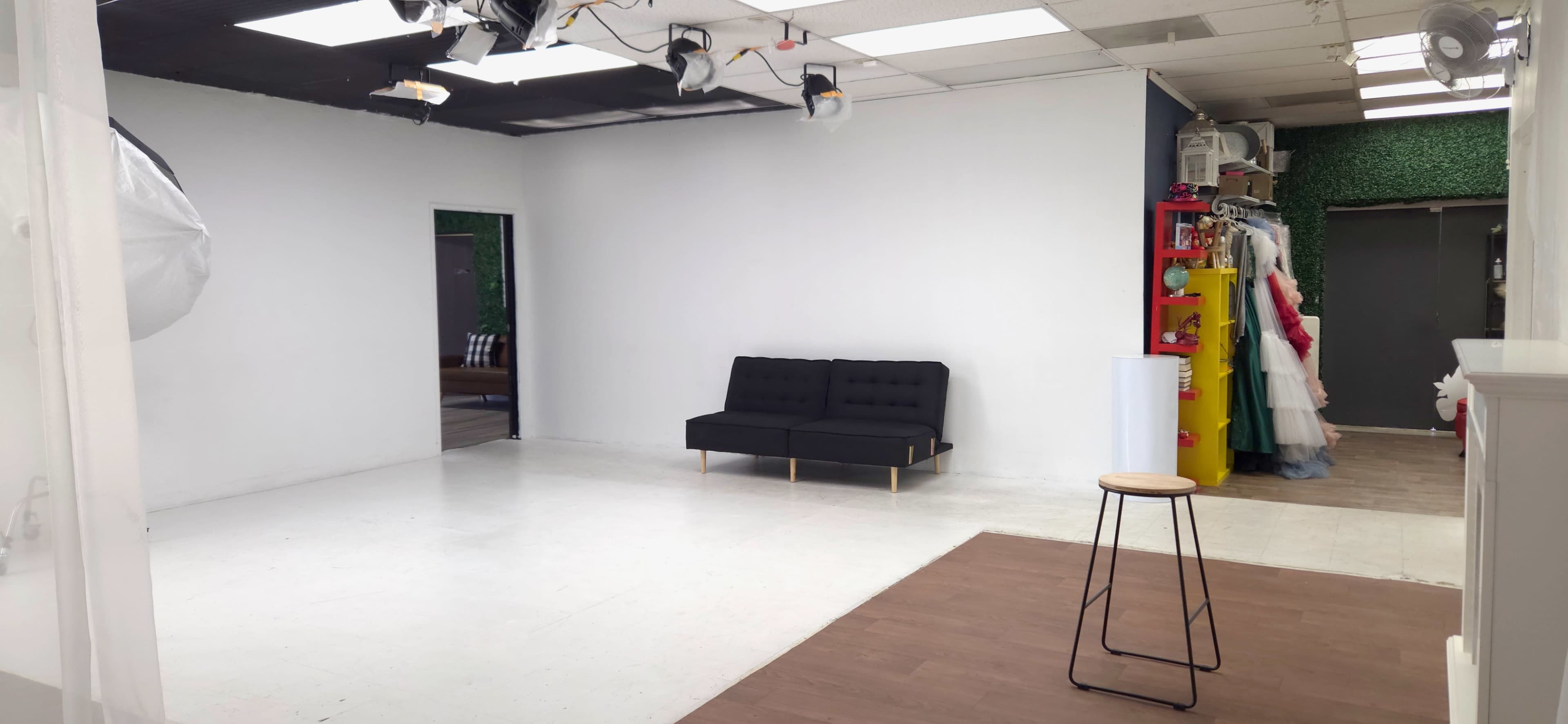 The image shows a minimalist studio space with a black sofa, a wooden stool, and colorful props lined along one wall.