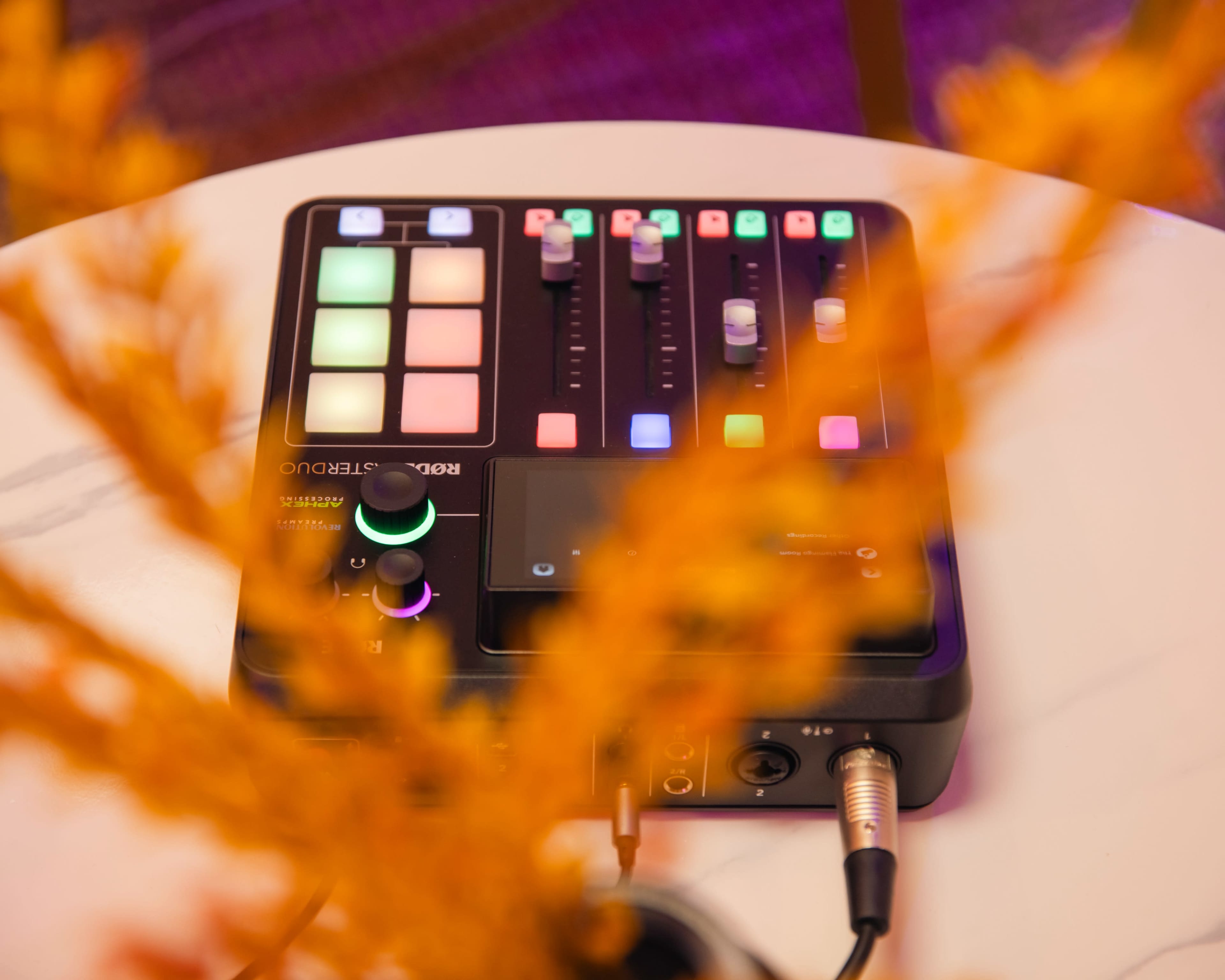 A black audio mixing device with colorful buttons and sliders is positioned on a white table, partially obscured by yellow foliage.