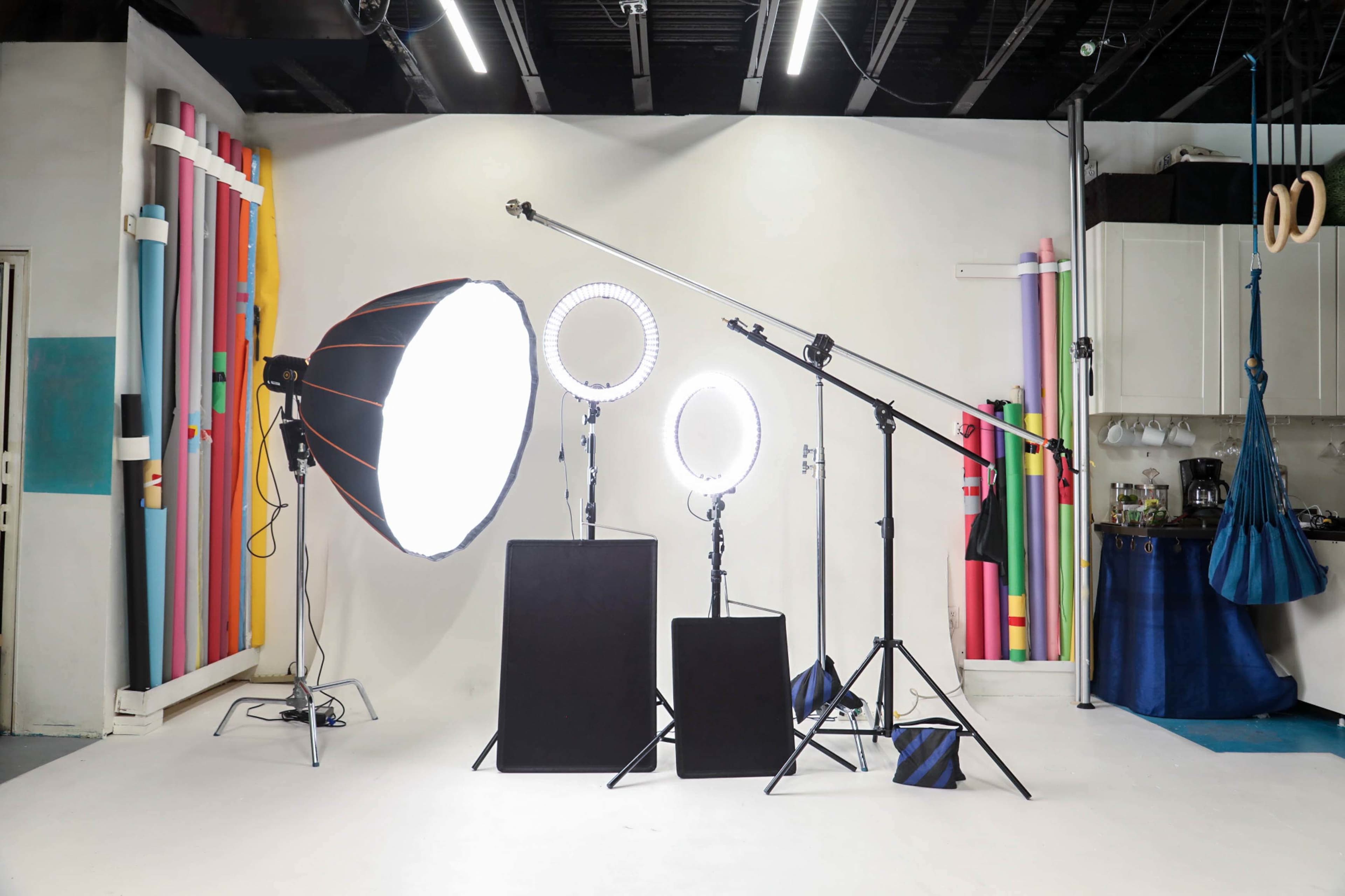 The image shows a photography studio setup featuring three lighting fixtures, two light stands, a backdrop, and colorful props arranged along the wall.